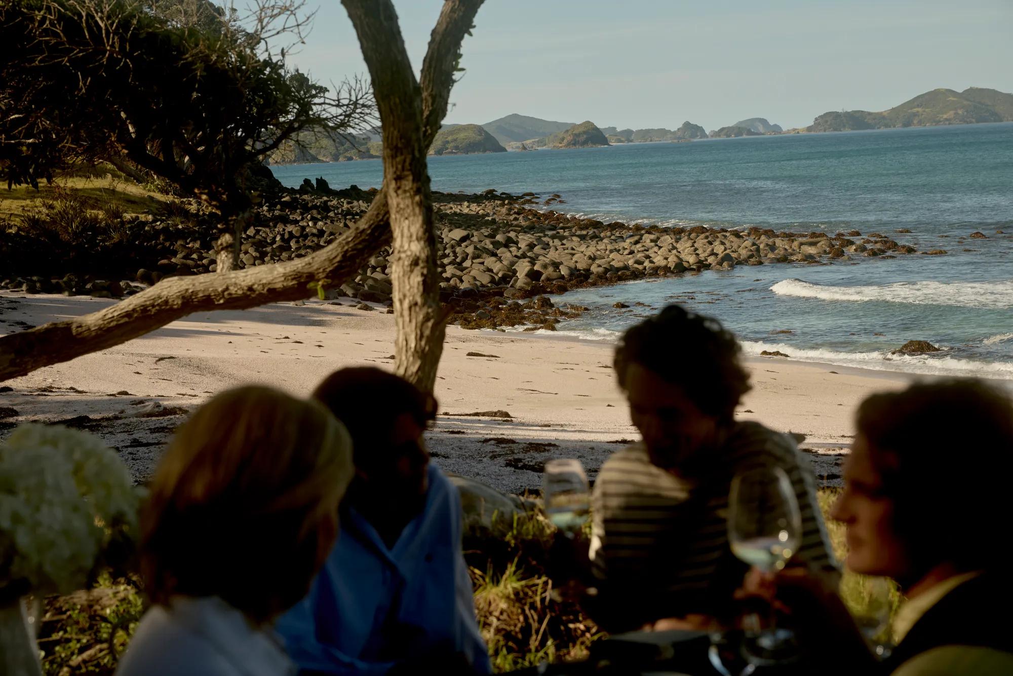Outdoor dining set up at Pink Beach including two wooden tables nicely set with fresh flowers, framed by Pohutukawa trees with the backdrop of the pink sand beach and the ocean.
