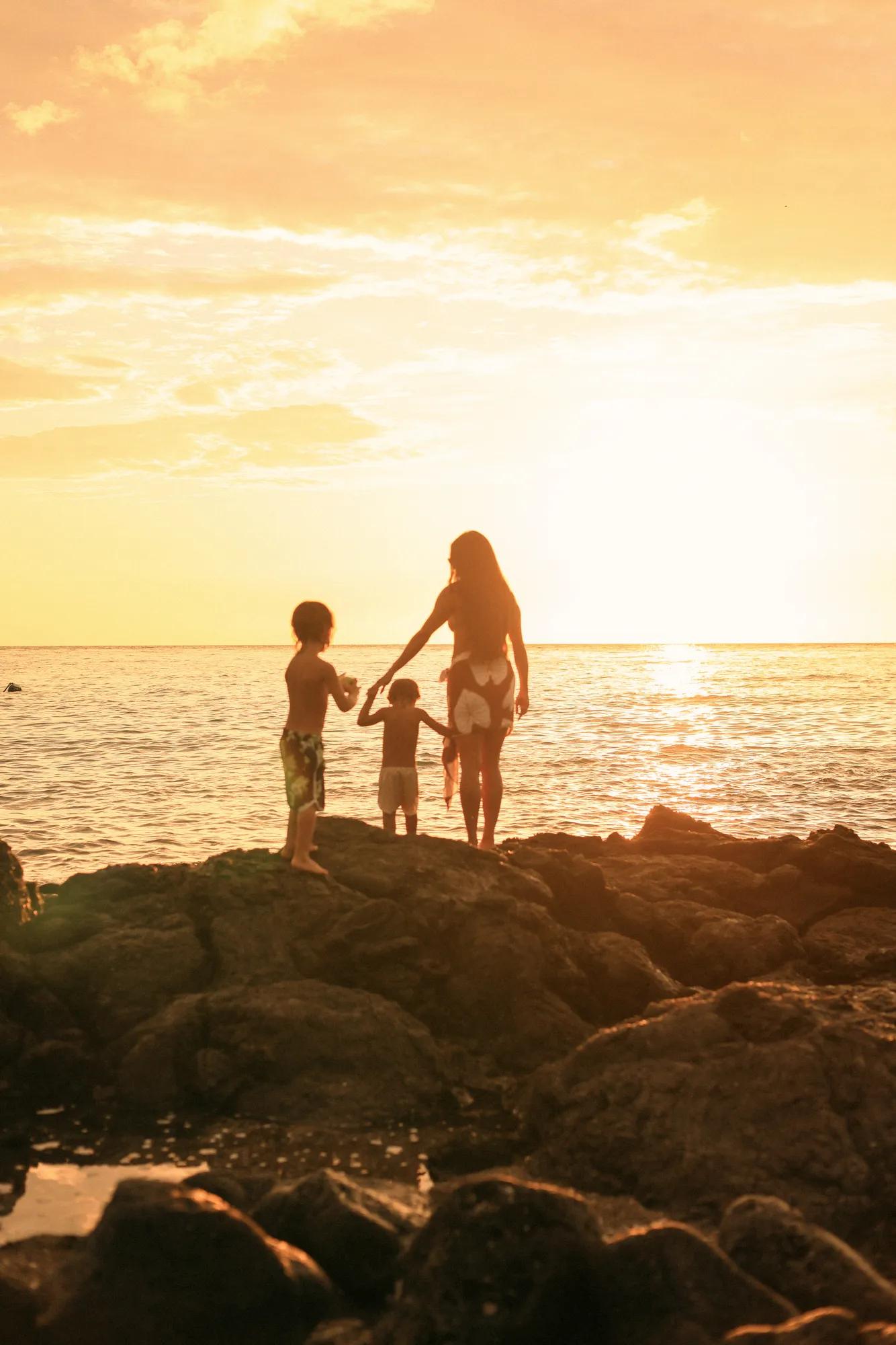 A mom and her two kids on lava rock shoreline at sunset