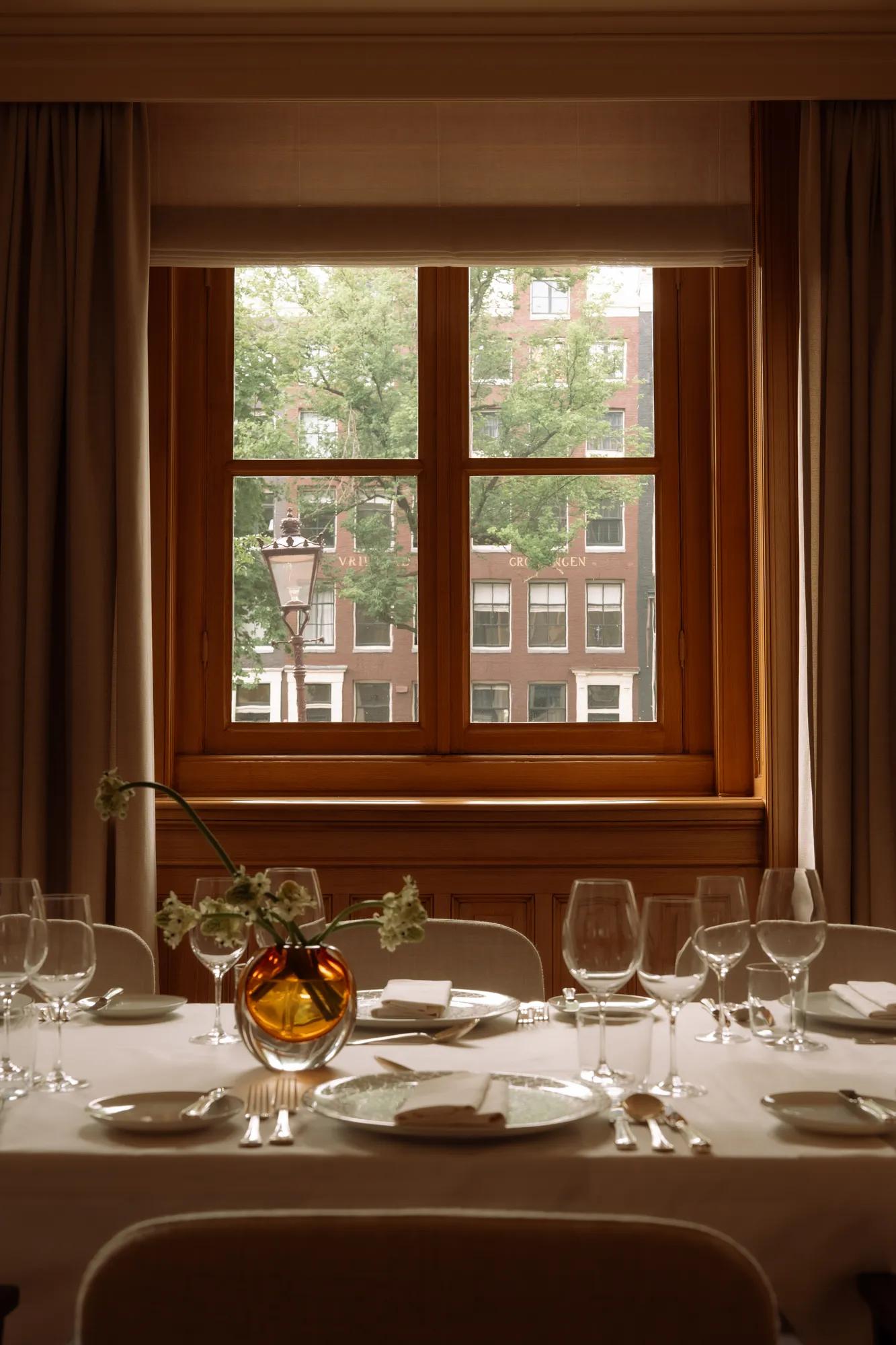 A table set up for dinner with glassware and white linen and a view of canal houses, trees and a traditional lamp post. 