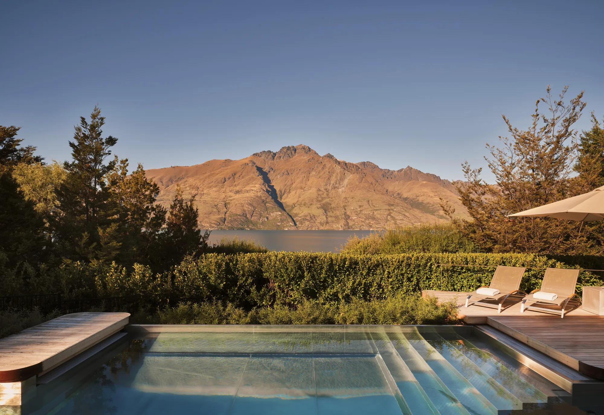 Front on view of an outdoor pool at a luxury lodge with sun loungers and views out to Lake Whakatipu and the surrounding mountains.