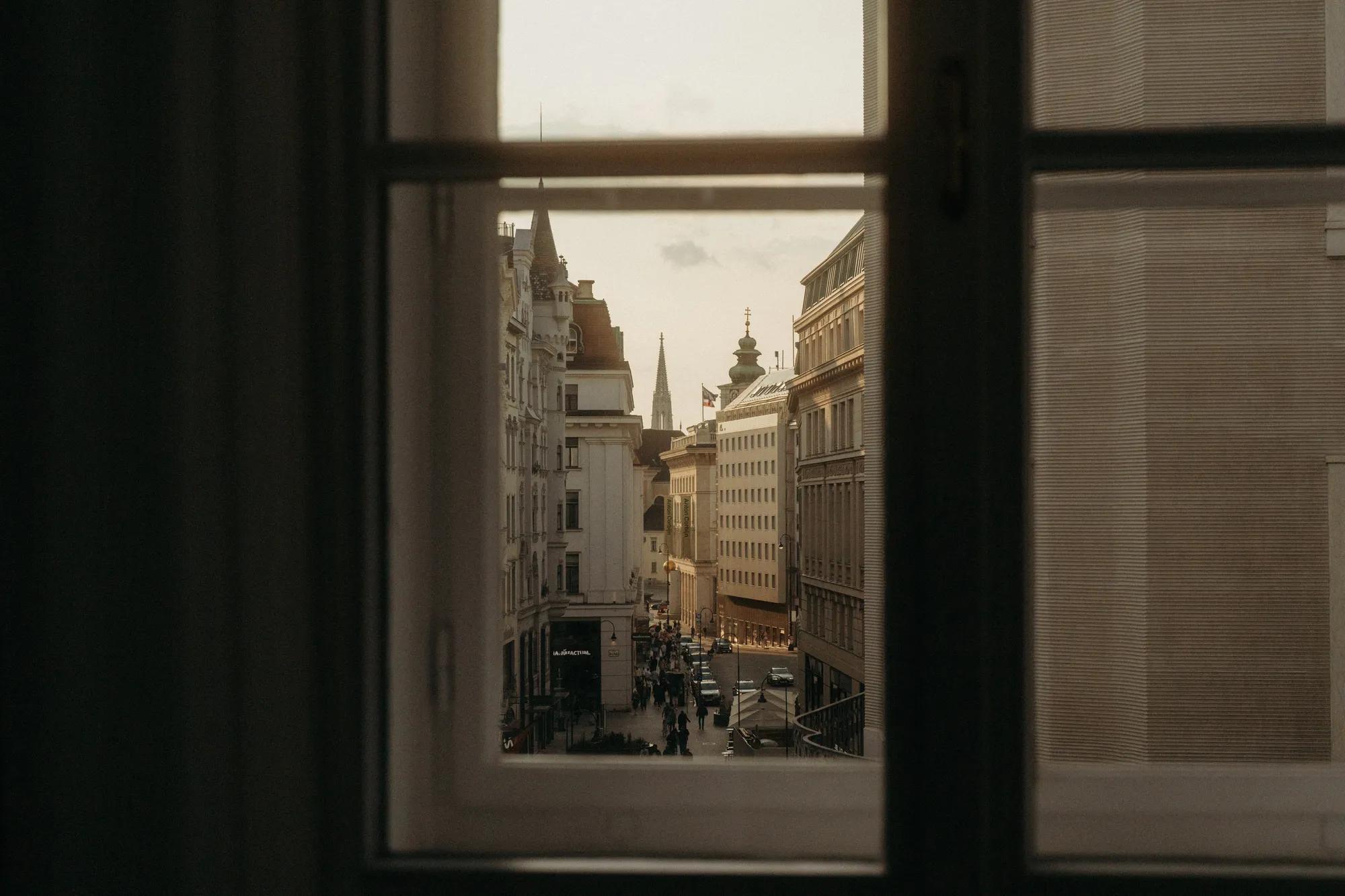 View of Vienna’s historic city center framed through a window, with bustling streets and church spires at dusk. "