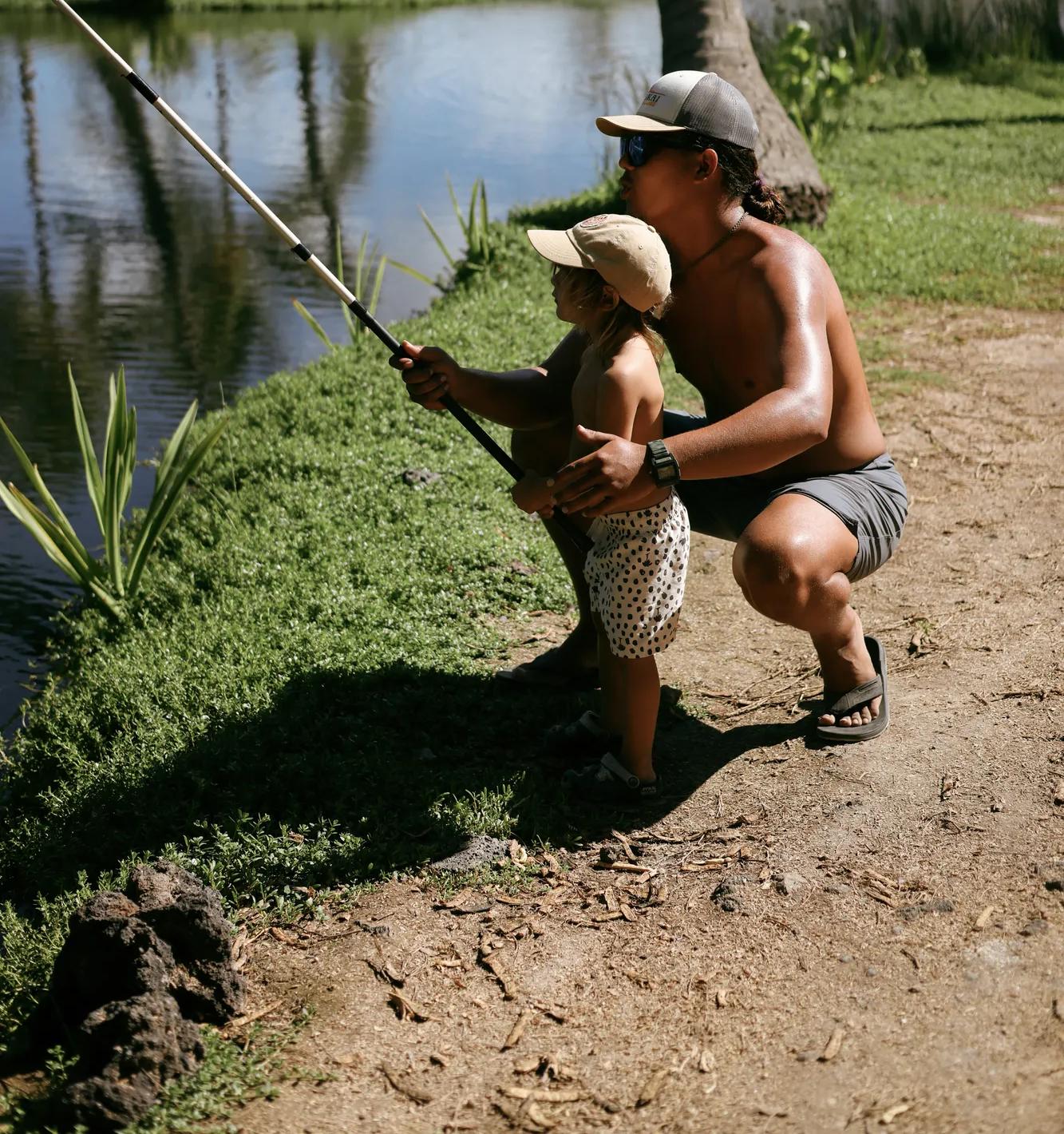 Man helping small boy fish in a pond with palm trees in the background.  