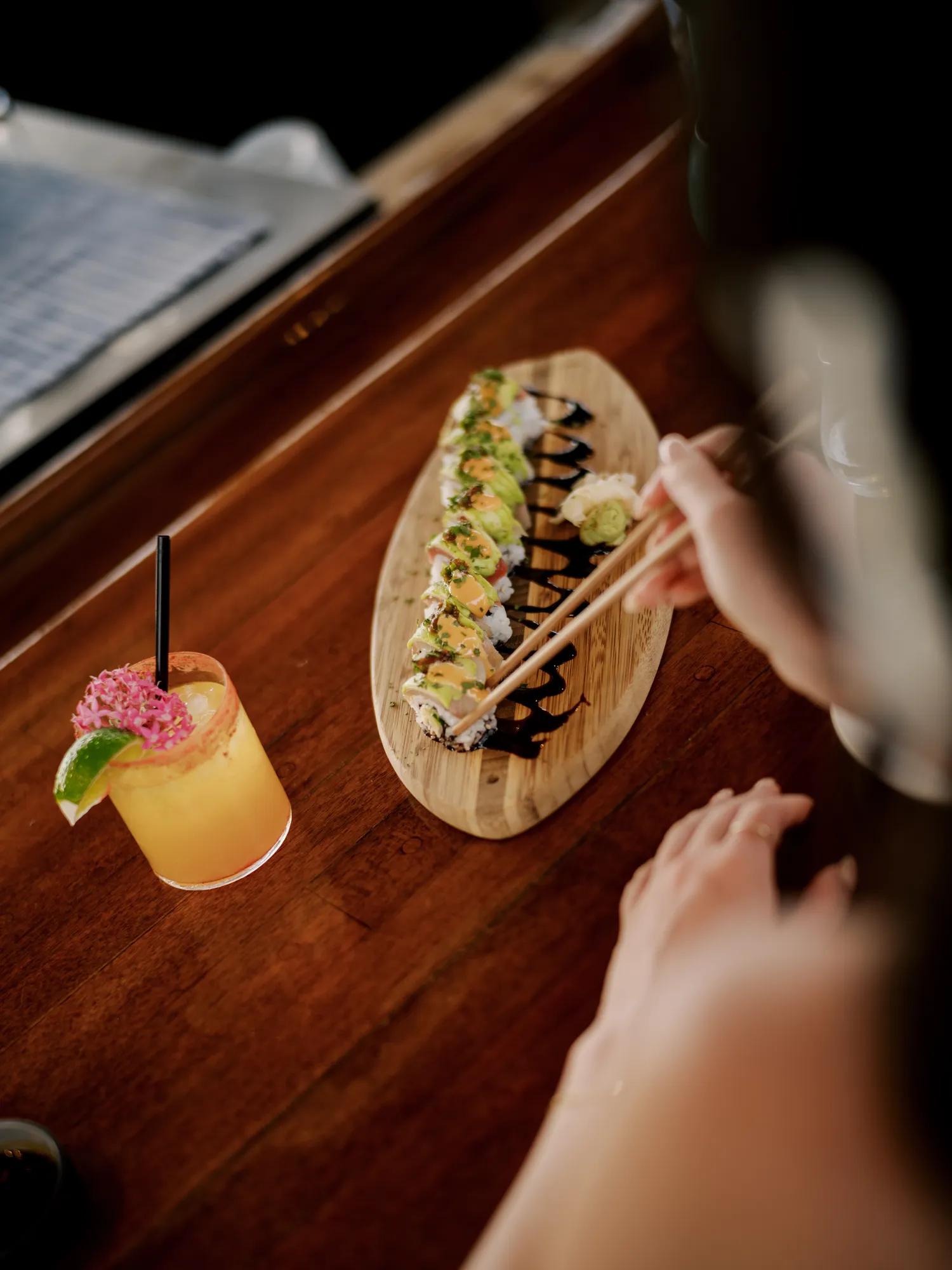 Person holding chopsticks picking up a piece of sushi with cocktail on the side