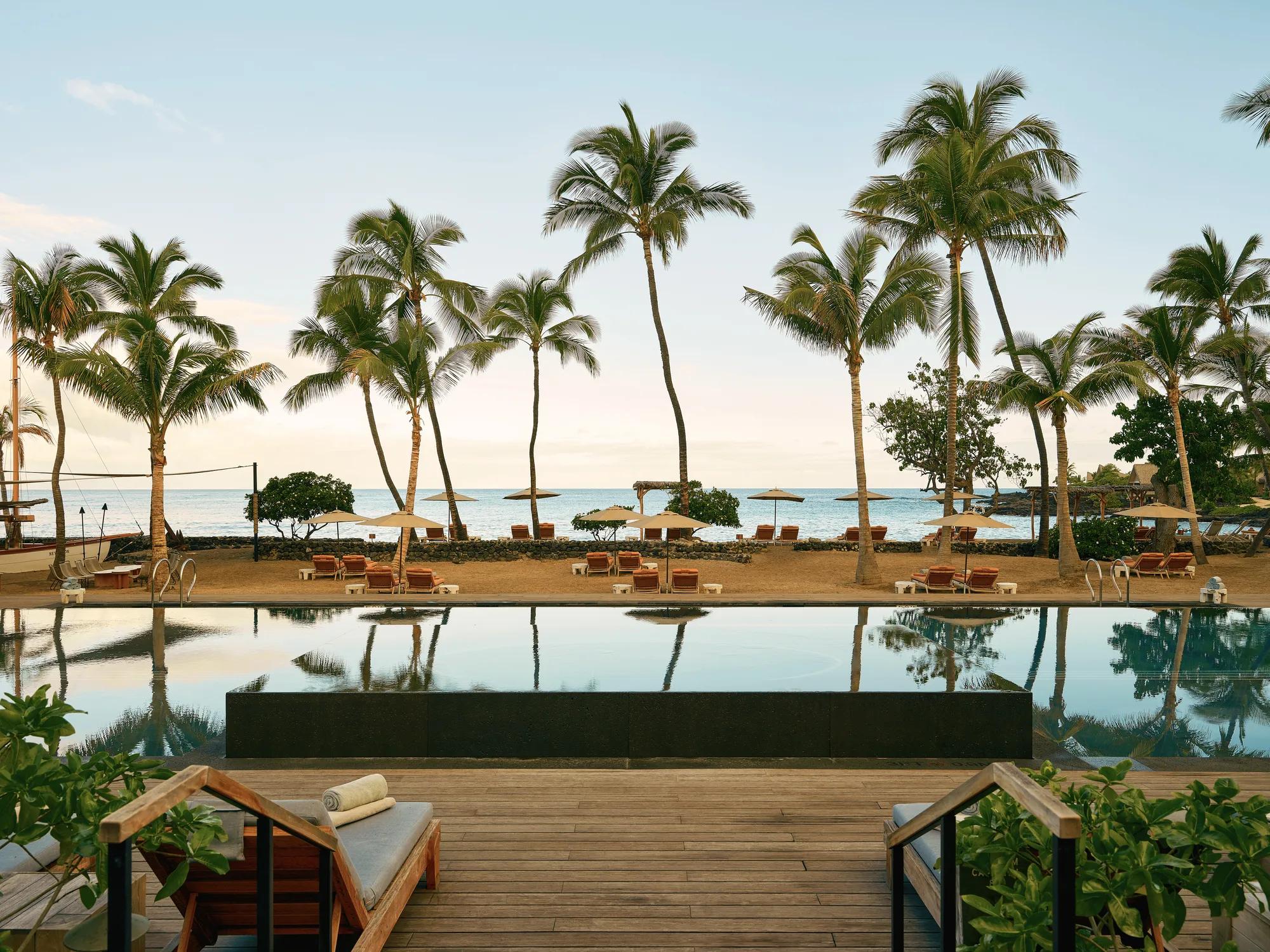 Infitnity hot tub into infinity pool overlooking the ocean with beach chairs spread out accross the shore and palms towering above the sand and pool.