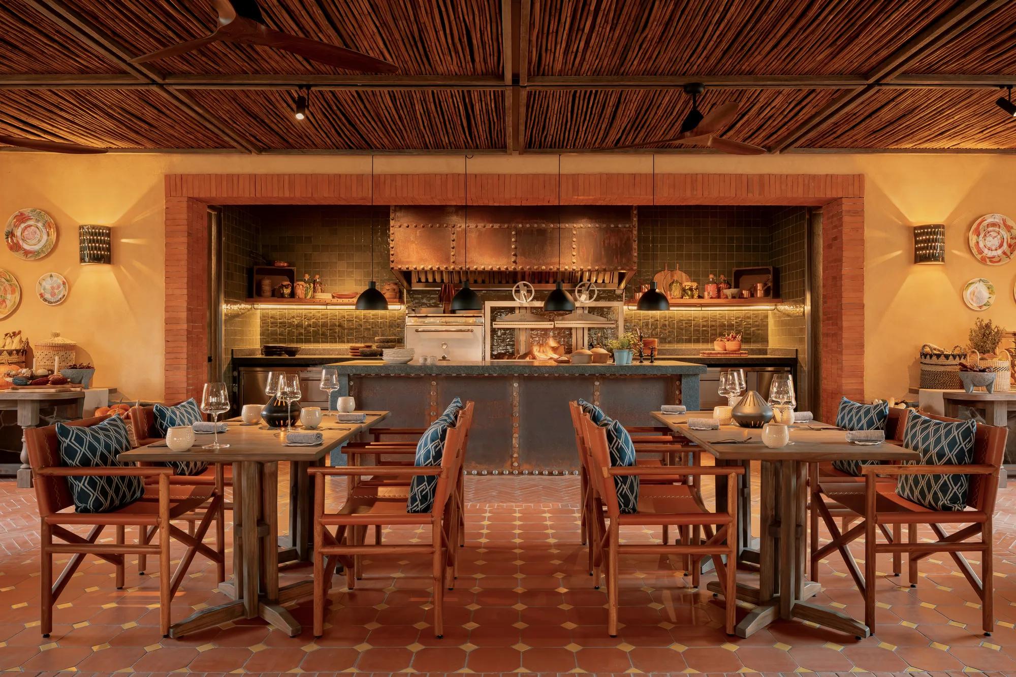 Open kitchen at Pirules Garden Kitchen with flames rising from the grill, and two tables set for four with blue-cushioned chairs in a lively garden setting.