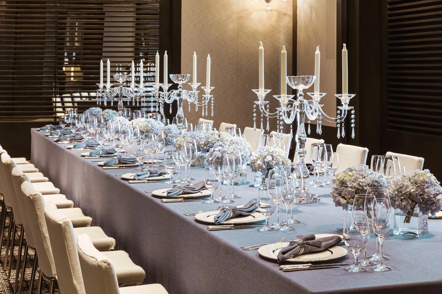 An elegantly set dining table with a light blue tablecloth, crystal candelabras, blue hydrangea centerpieces, and white upholstered chairs arranged for a formal event.