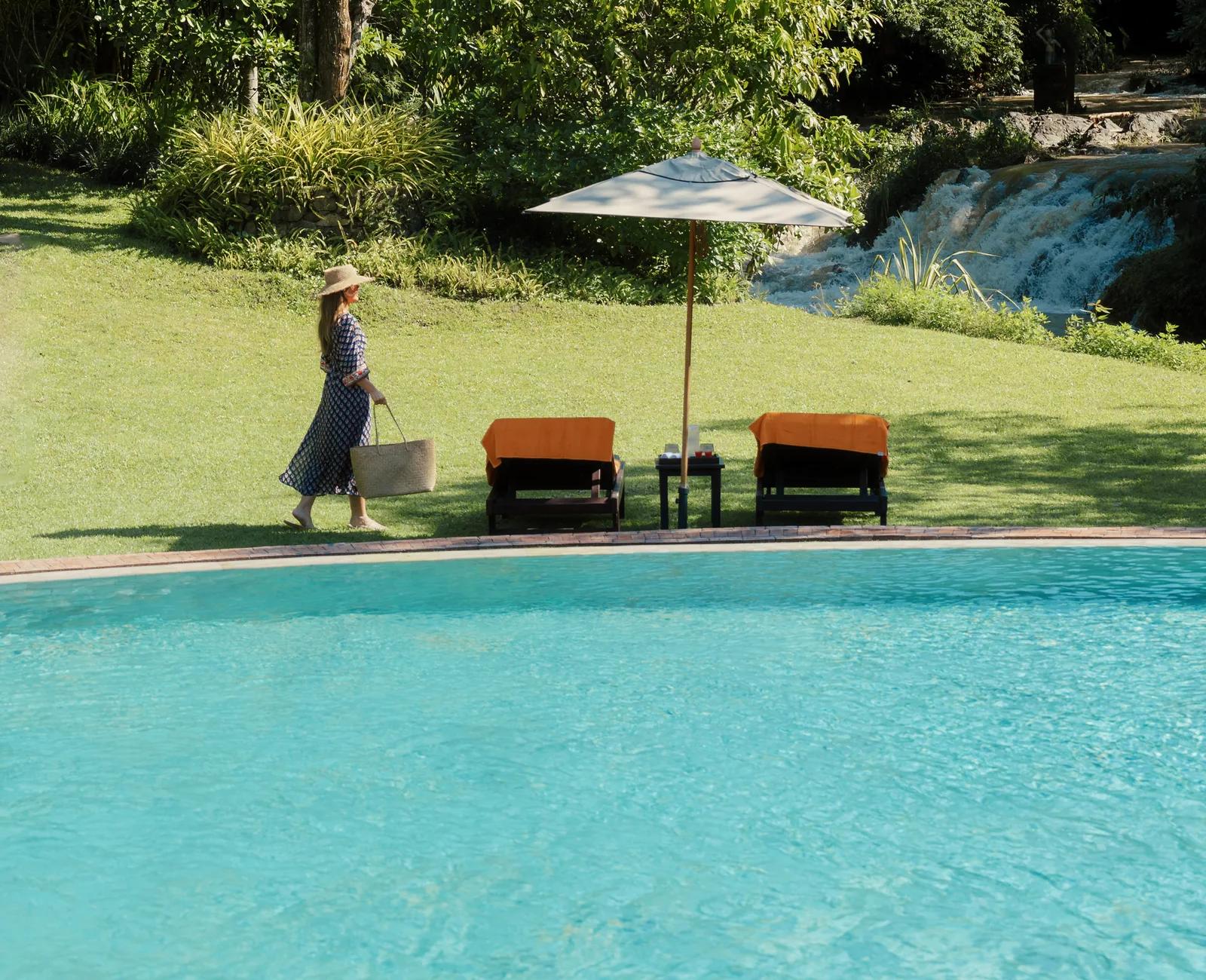 A guest walking by a lawn with a pool and orange loungers beside a waterfall backdrop.