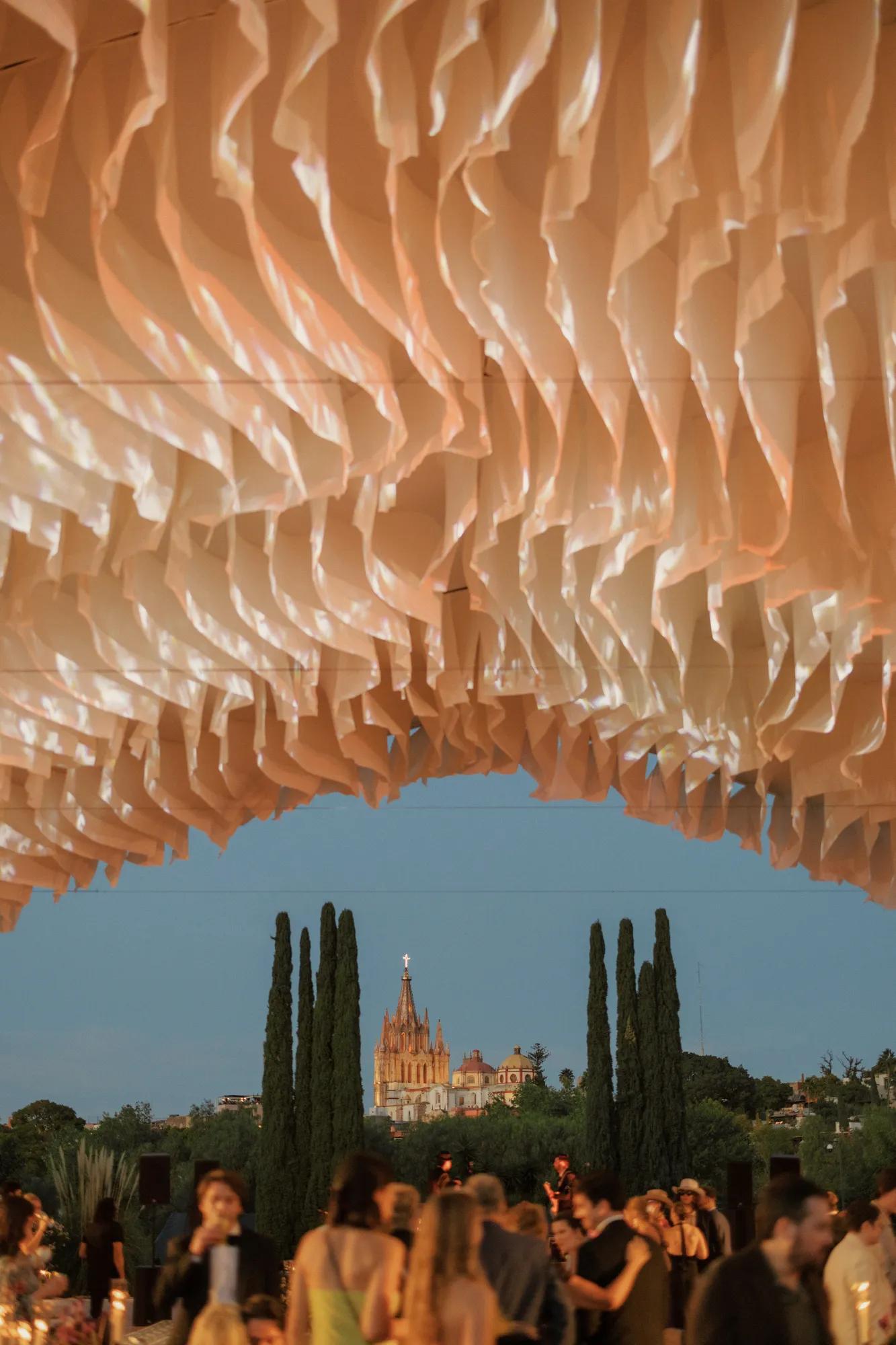 Event setup at Jardín Rosewood with guests gathered under a draped canopy, set on a garden lawn with the Parroquia de San Miguel Arcángel in the background.