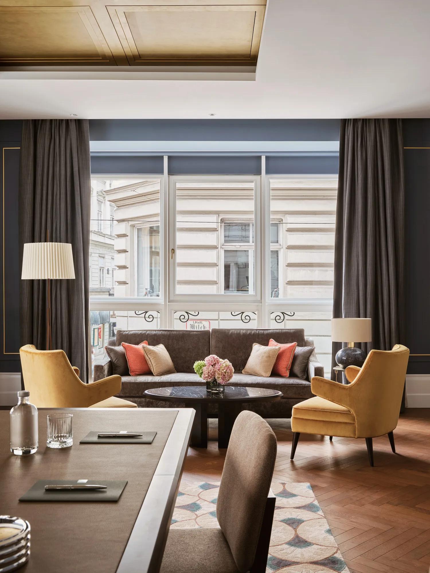 Elegant event space at Rosewood Vienna featuring a stylish seating area with plush armchairs, a sofa, and a central table adorned with fresh flowers, set against large windows.