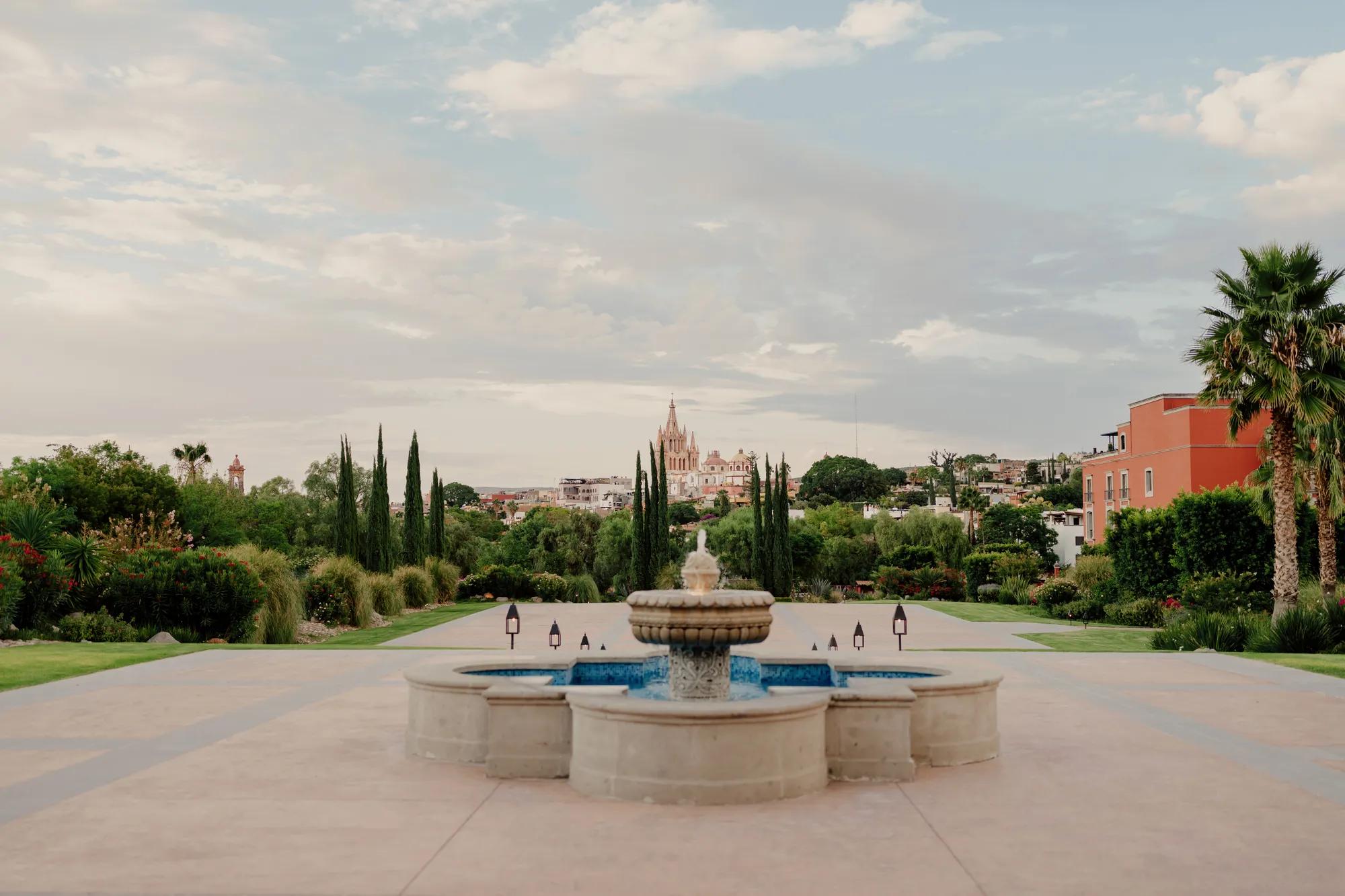 Stone fountain in the foreground with a spacious garden behind it, framed by greenery and a distant view of the Parroquia de San Miguel Arcángel.