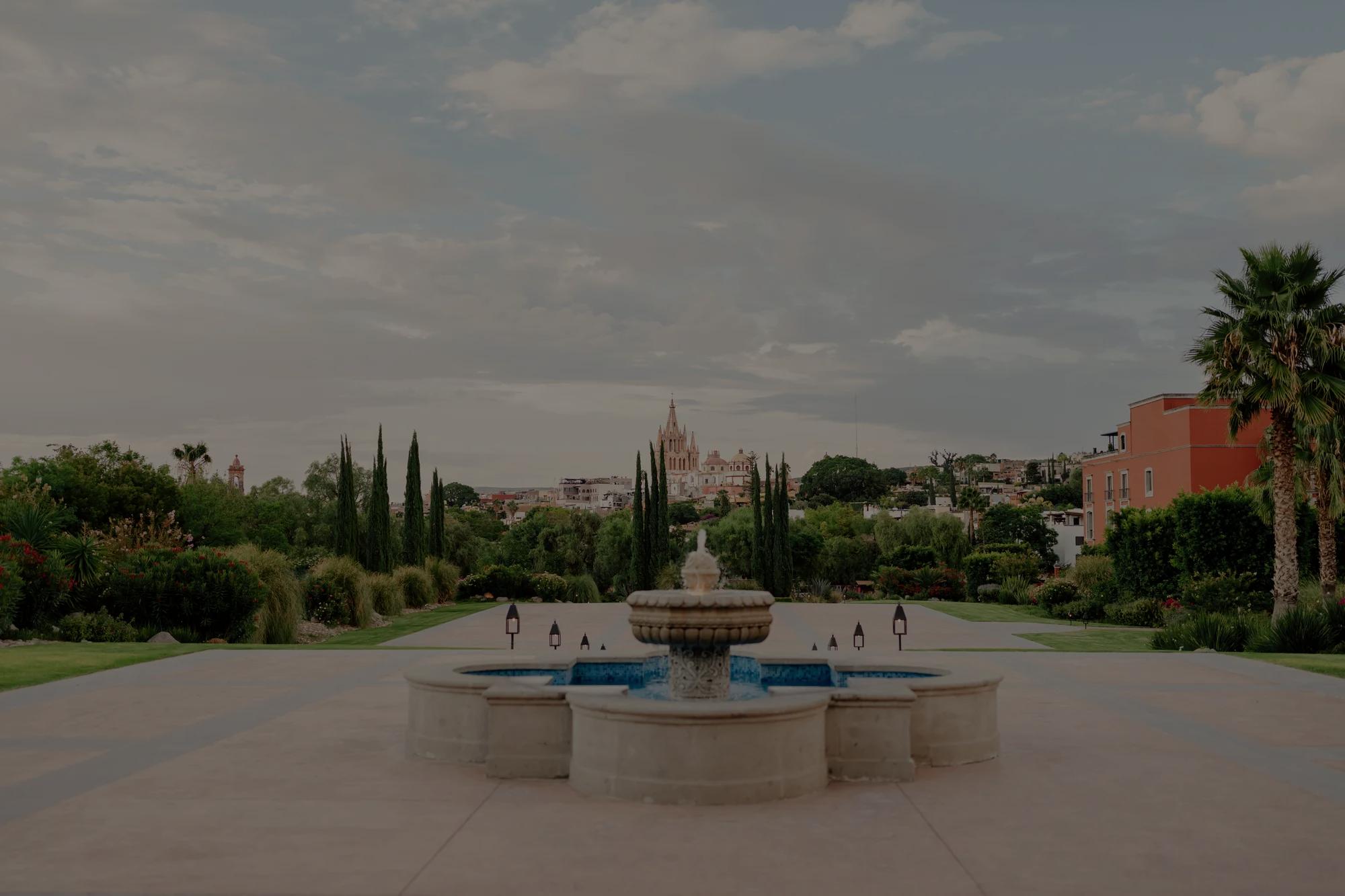 Stone fountain at Jardín Rosewood, nestled between two esplanades, with lush gardens and the Parroquia de San Miguel Arcángel in the background.