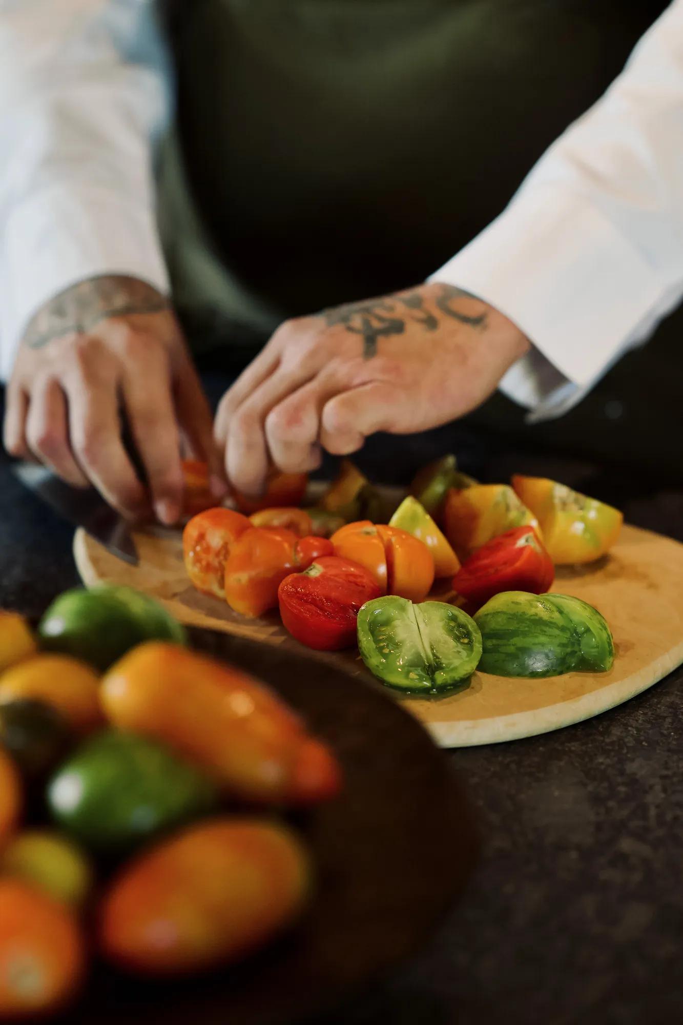 Wooden cutting board with halved colorful tomatoes, a bowl filled with more tomatoes in front, and a chef's arms slicing the tomatoes.