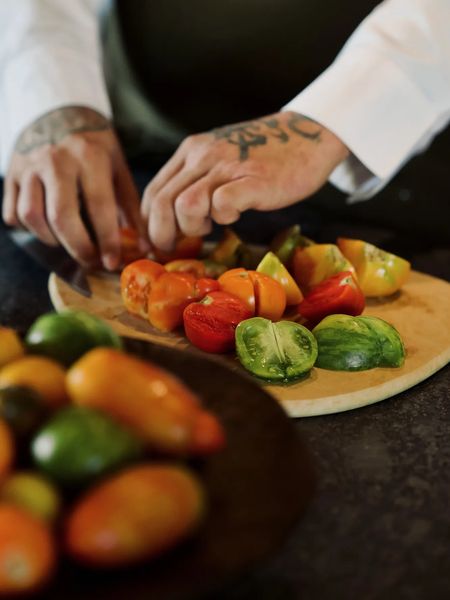 Wooden cutting board with halved colorful tomatoes, a bowl filled with more tomatoes in front, and a chef's arms slicing the tomatoes.