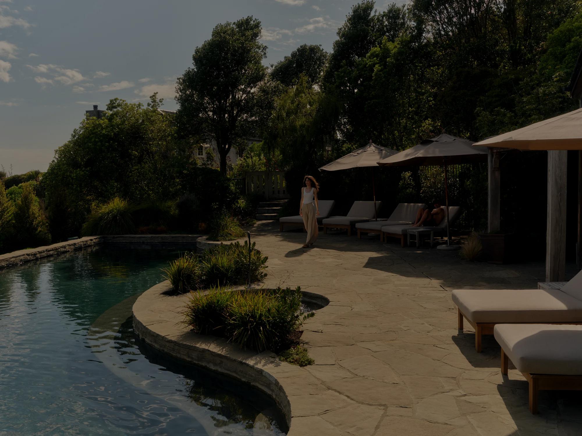 Couple relax by an outdoor pool, a woman walks towards the pool while her partner lounges on a deck chair shaded by an umbrella.
