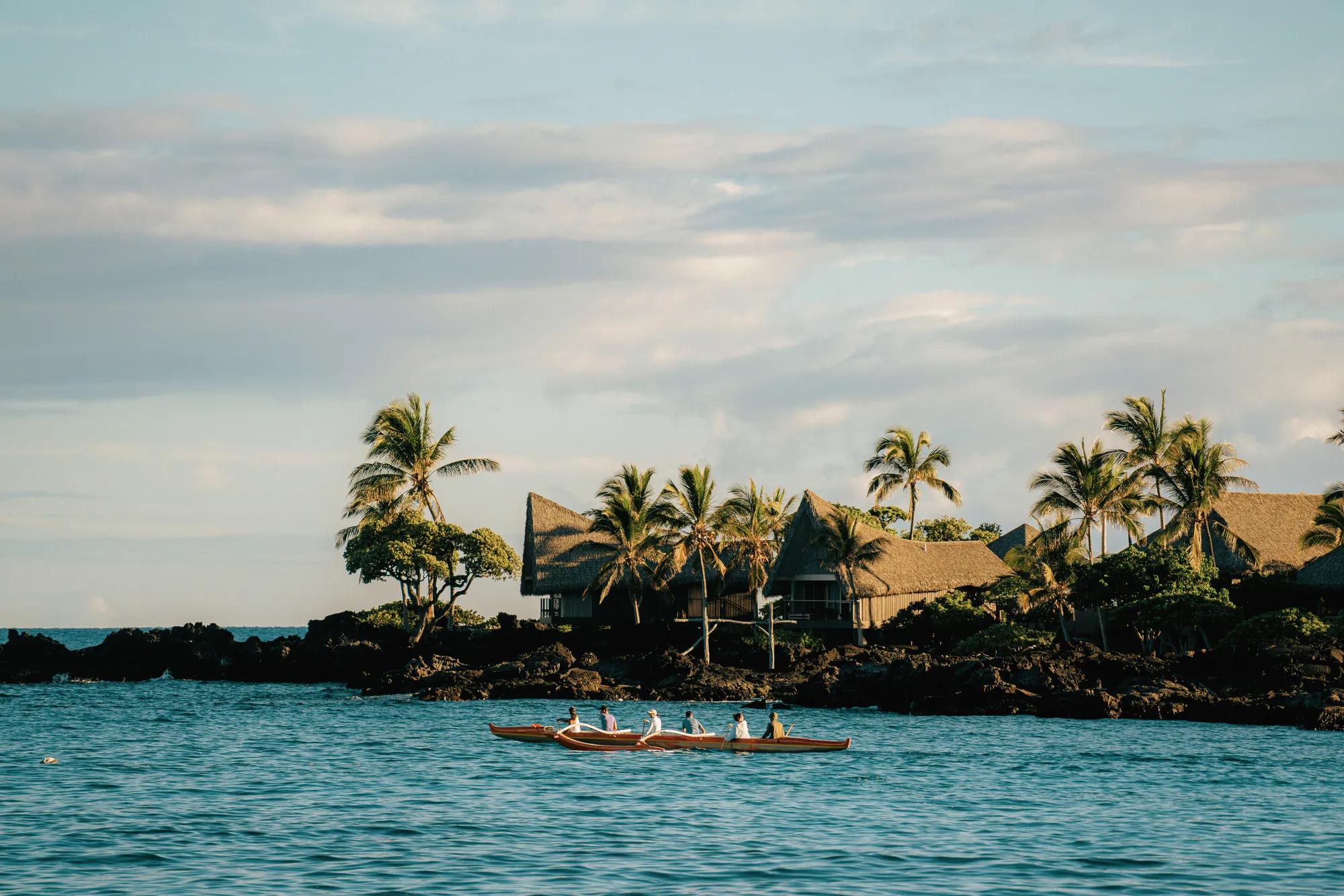 Traditional outrigger canoe on water with rooms and lava rock coastline in the distance