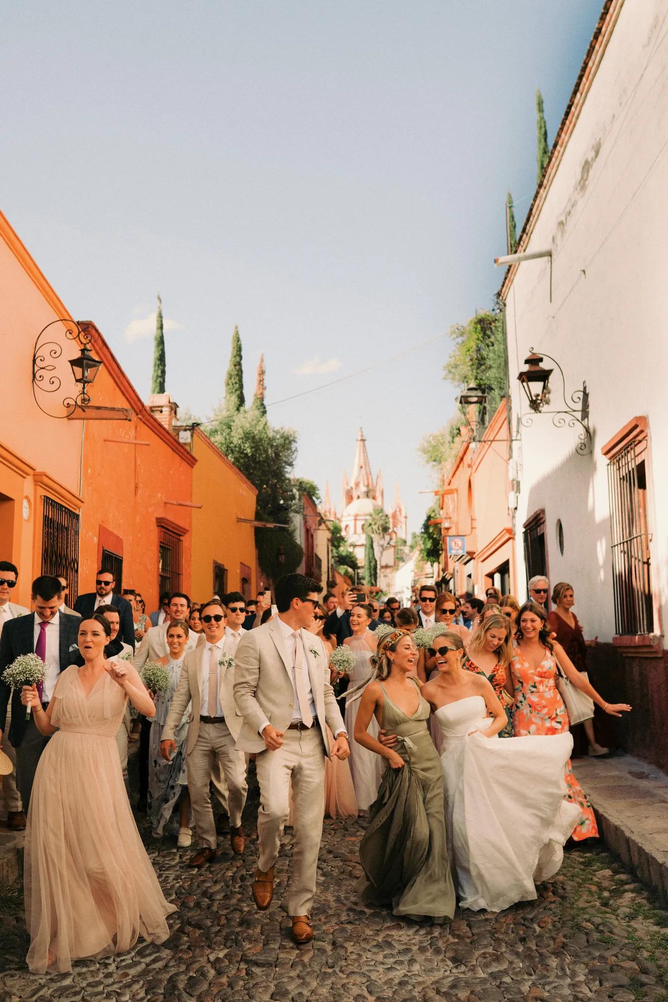 Bride and groom walking through the cobblestone streets of San Miguel de Allende during a traditional callejoneada, followed by guests celebrating together.