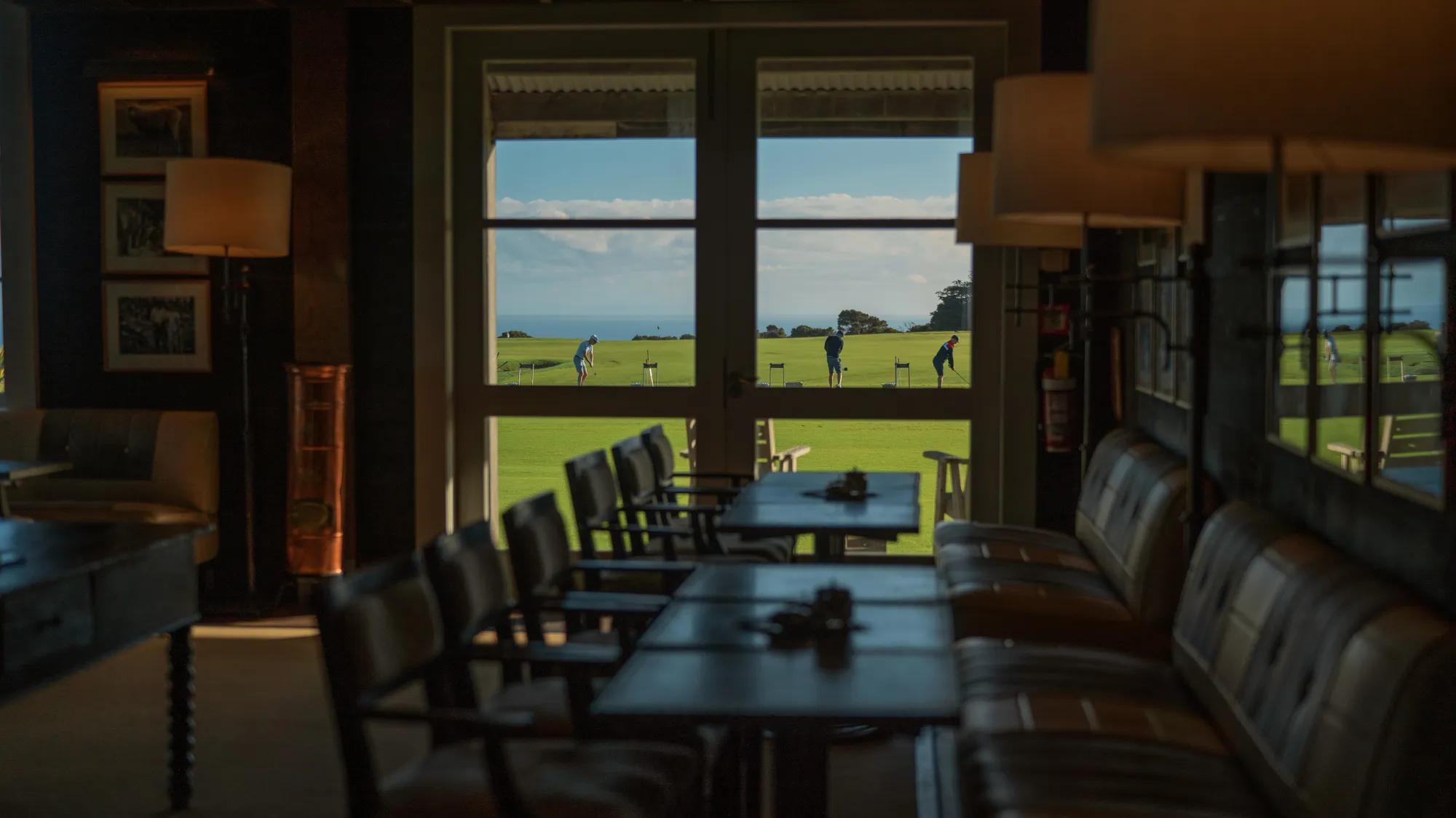 A darkened dining room in the Clubhouse with a view through the window to players practicing on the driving range outside. 