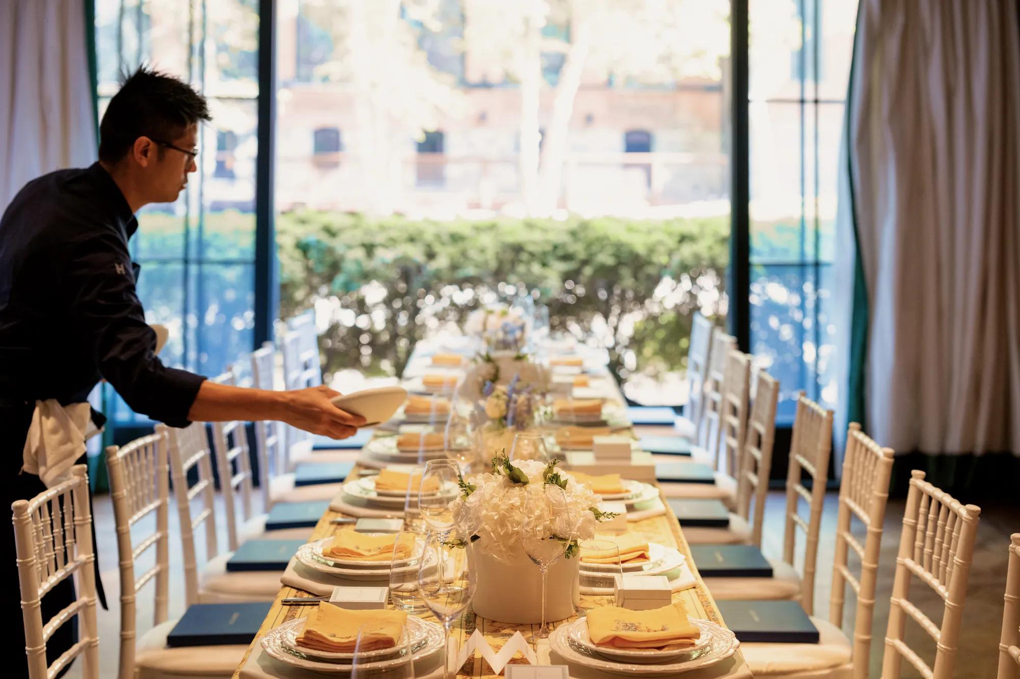 Event table at Rosewood Washington D.C. with final touches being done by a chef. Features yellow linen, white flowers, and white chairs.