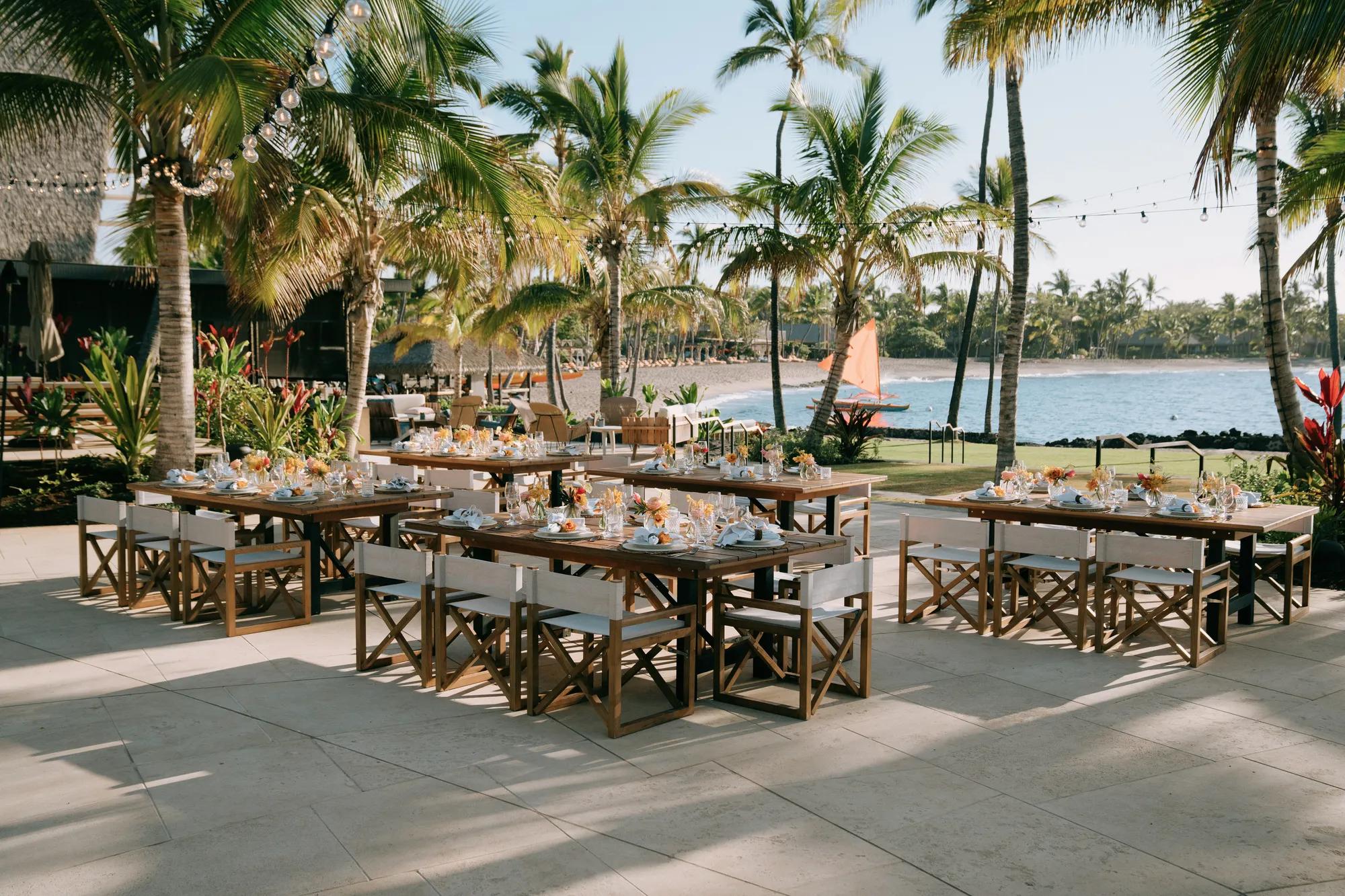 Tables set up against the backdrop of the ocean with flowers on each table and an orange sailing canoe beached in the distance