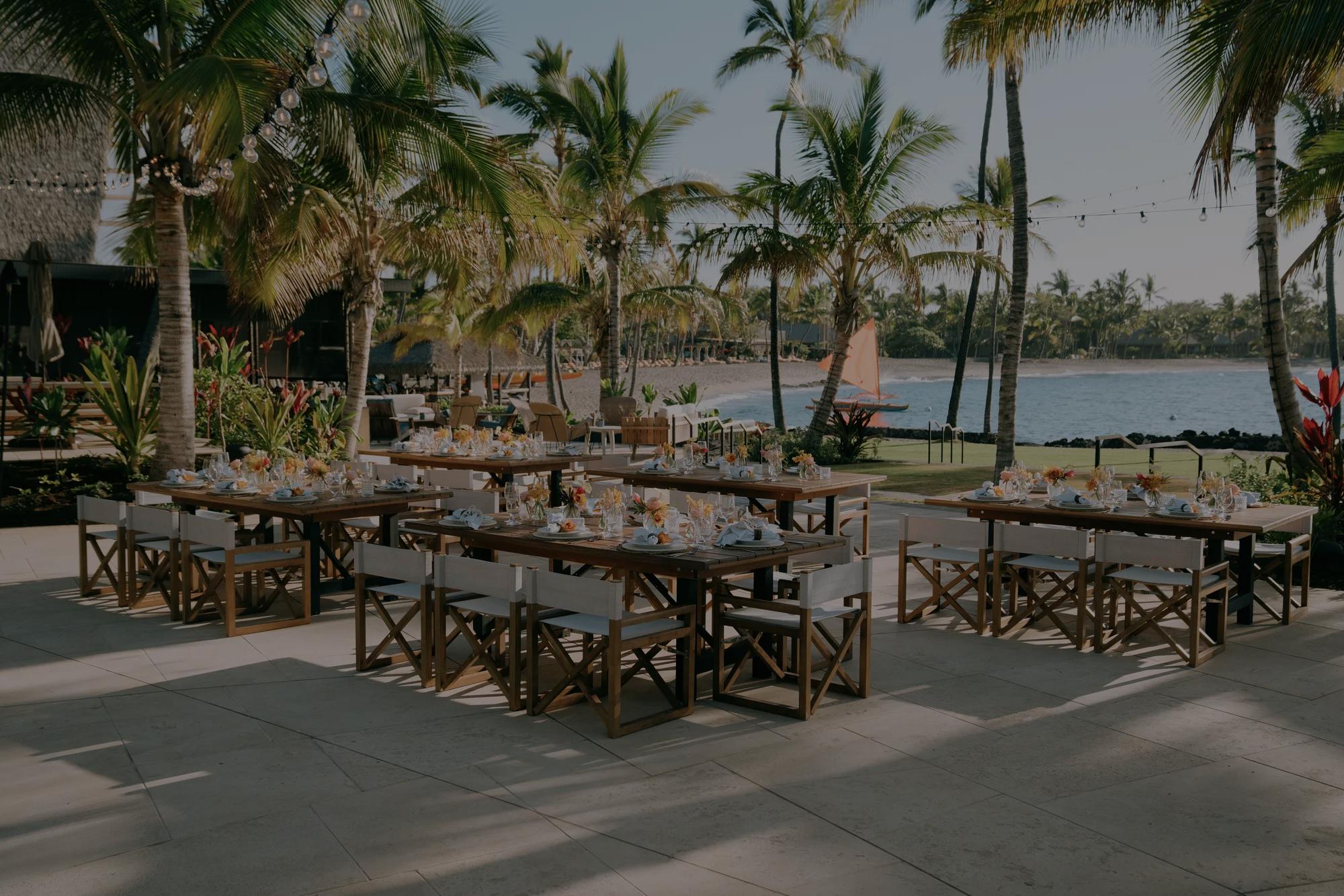 Tables and charis set for an event on a deck wtih paln trees and the beach and ocean in the background. 