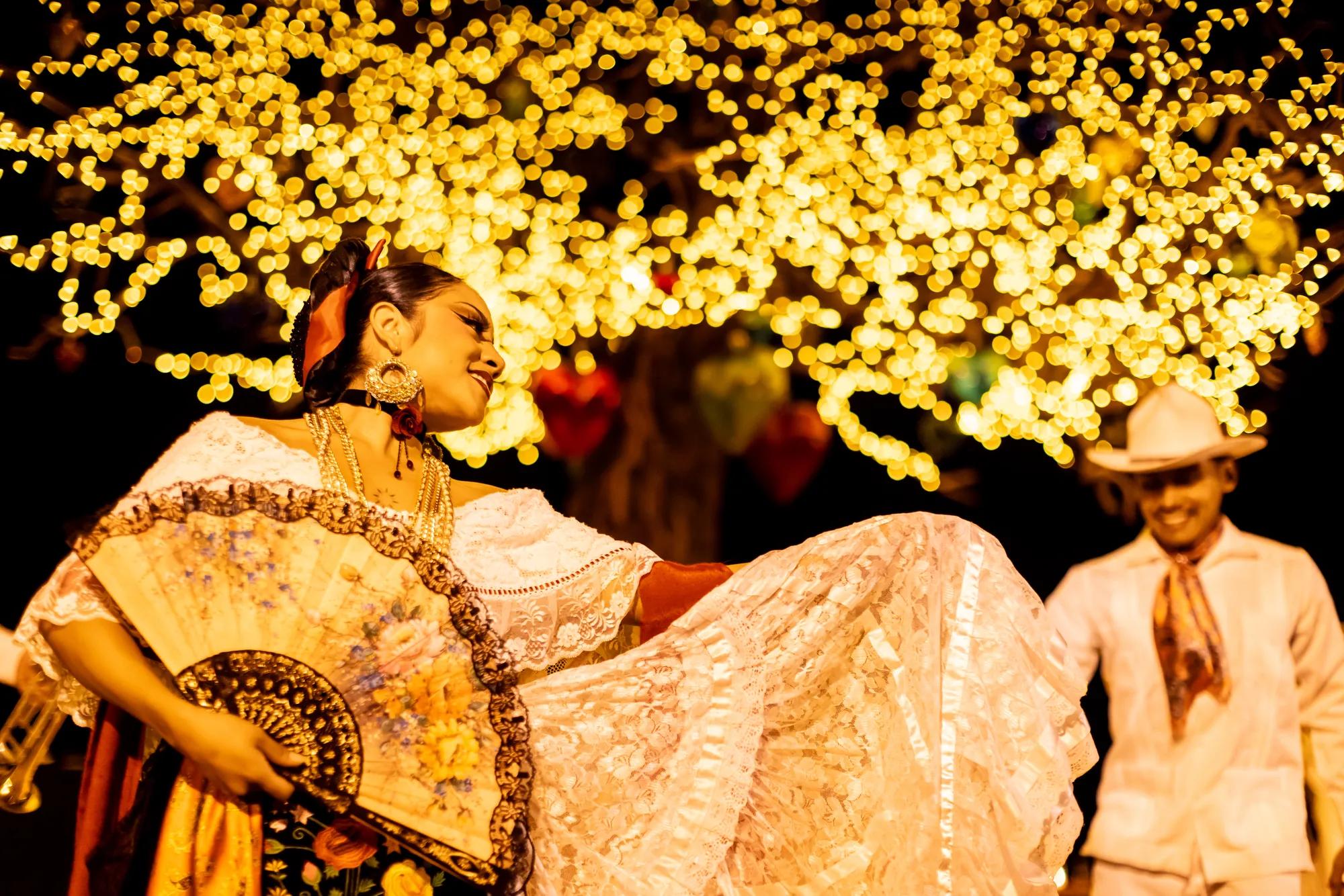 A woman in traditional folkloric dress dances gracefully, holding a decorative fan and lifting her lace skirt, under glowing golden lights. A man in a white outfit and hat stands smiling in the background, creating a festive Día de Muertos atmosphere.