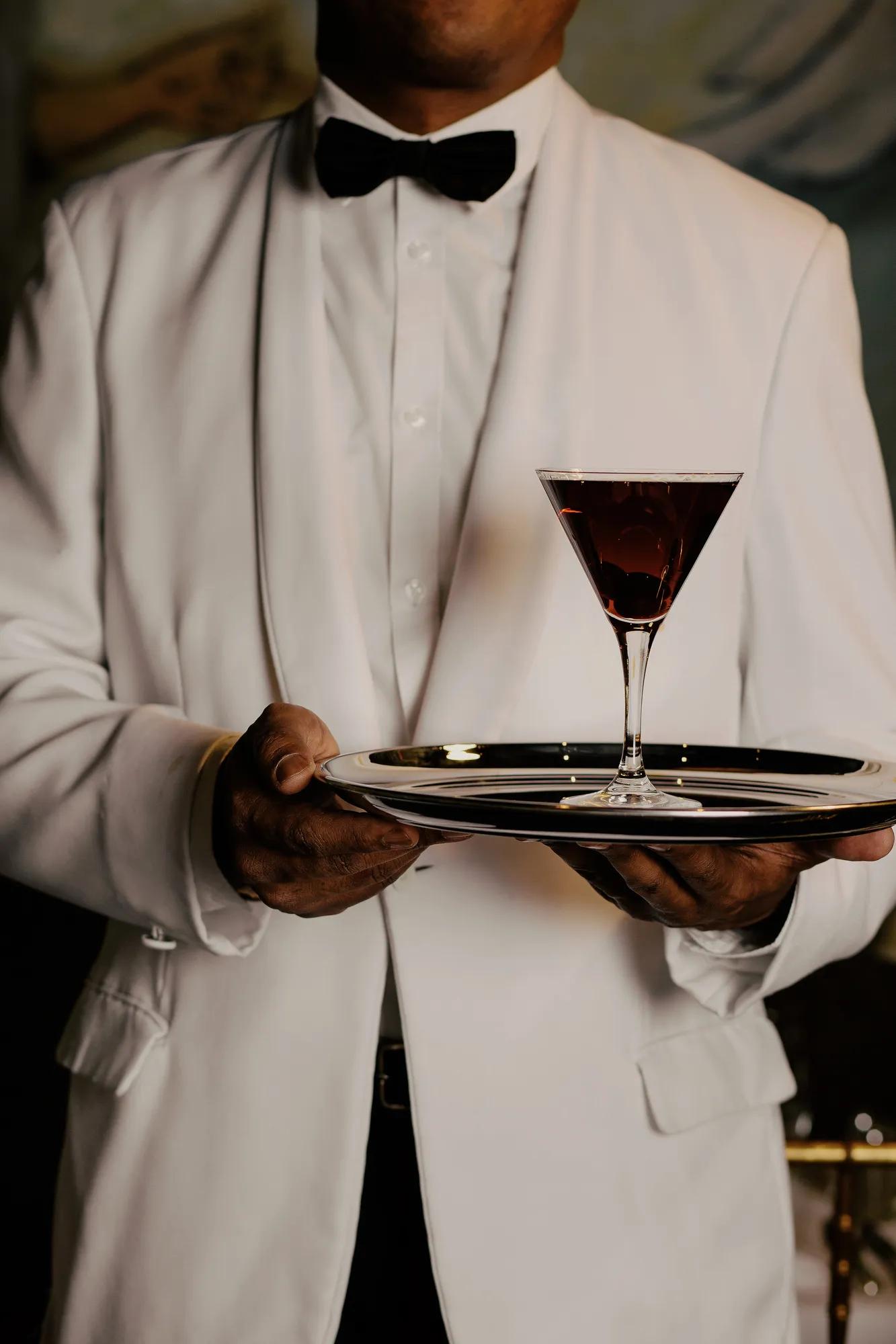 A server holding a custom cocktail in a white jacket and bowtie. 