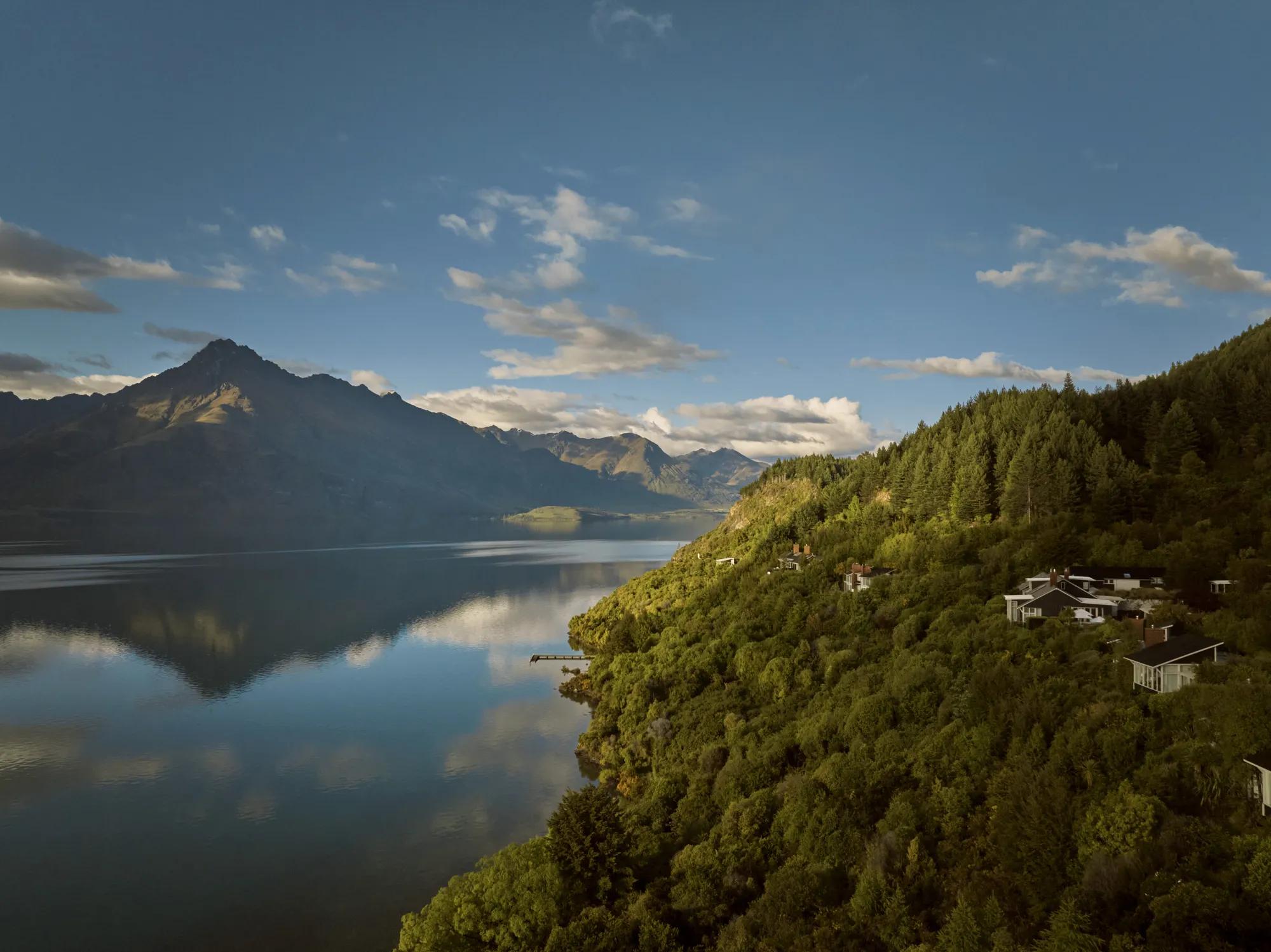 Aerial view of Rosewood Matakauri tucked into native bush, to the left the perfectly still Lake Wakatipu reflects the surrounding mountain ranges.