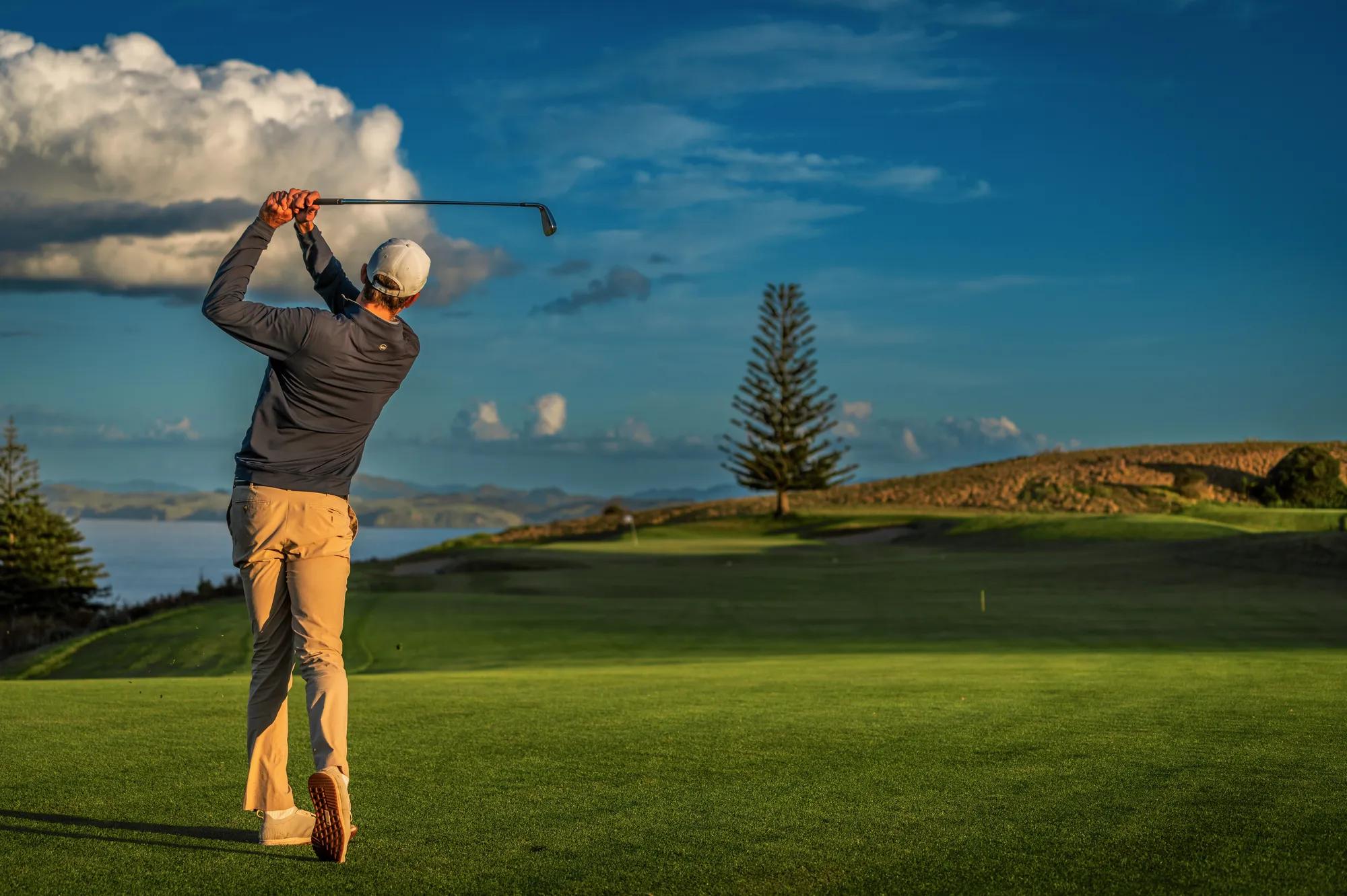 Man finishing a golf swing down a lush green fairway with the Pacific Ocean in the distance.