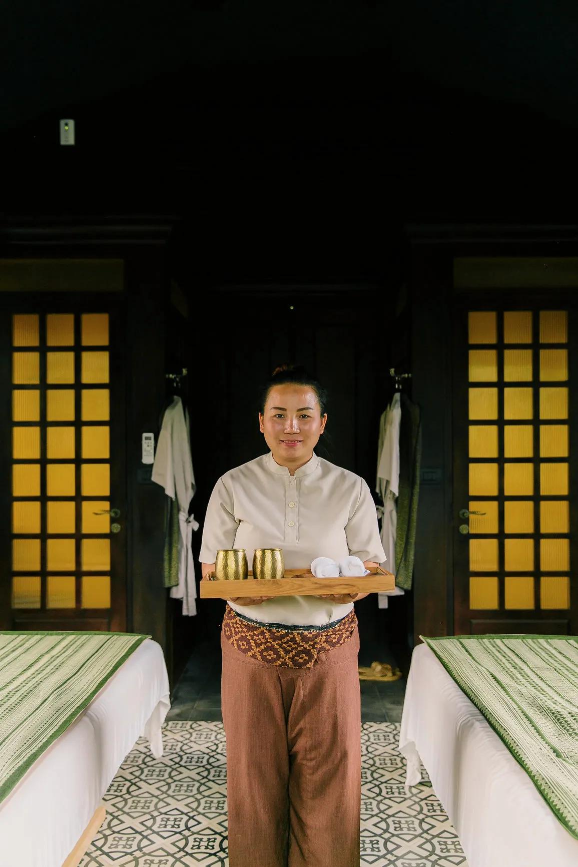 Spa attendant holding a wooden tray with gold cups and towels between massage tables.