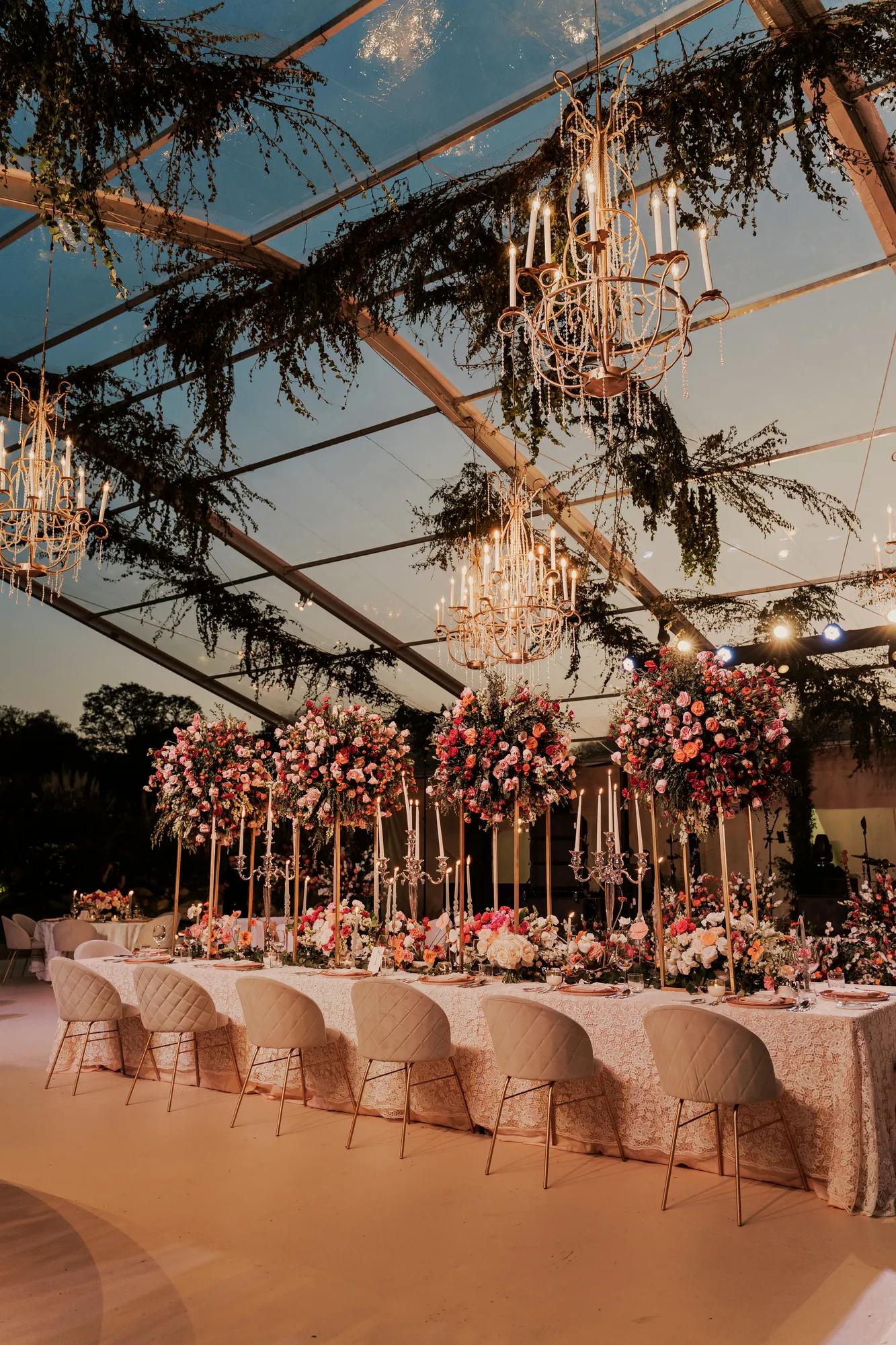 Outdoor wedding setup in soft pink tones, with matching table linens, chairs, and floral arrangements under a transparent canopy adorned with hanging greenery, all bathed in the warm light of sunset.