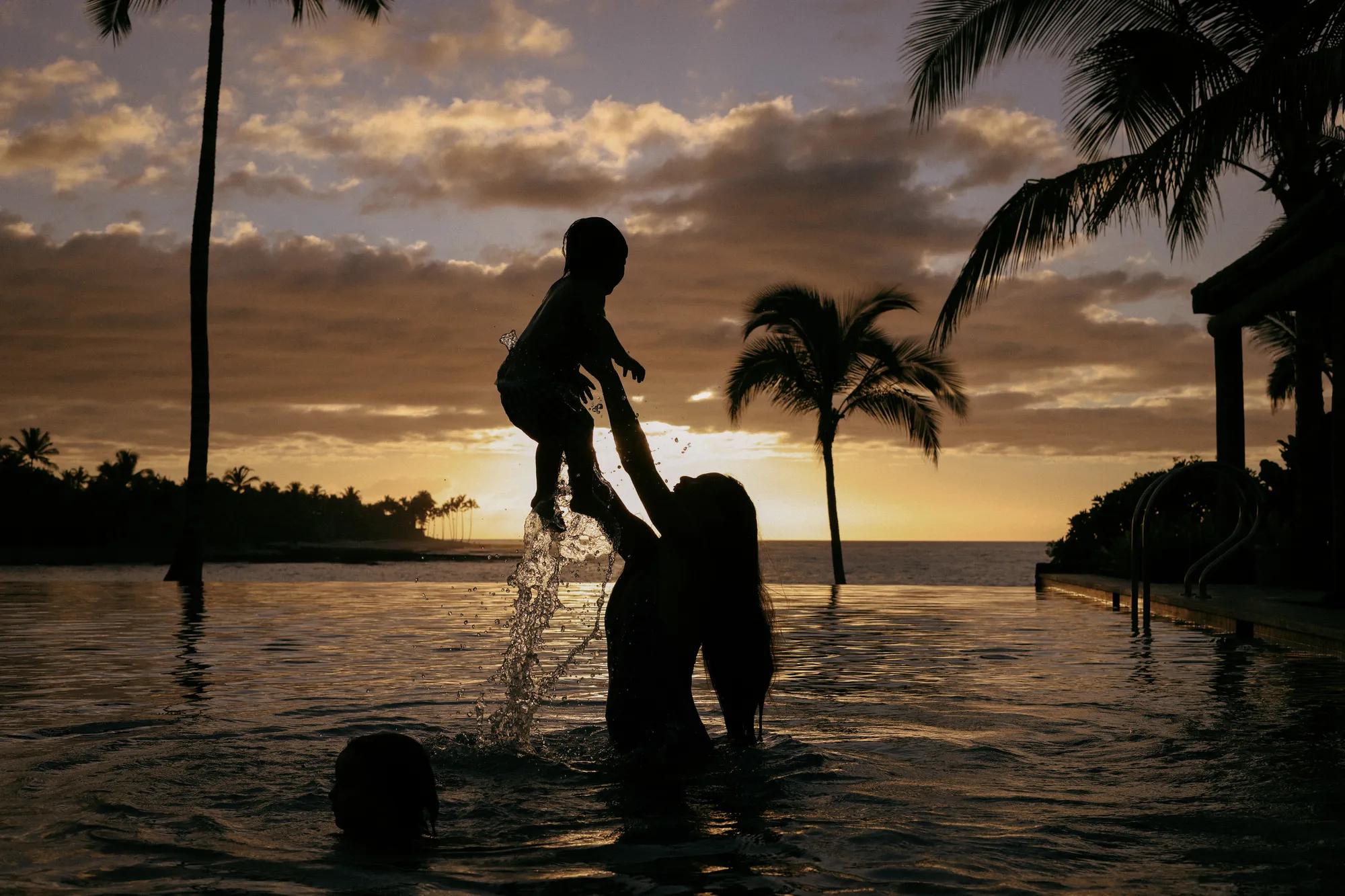 Mother throwing son in the air in a pool with palm trees in the distance. 