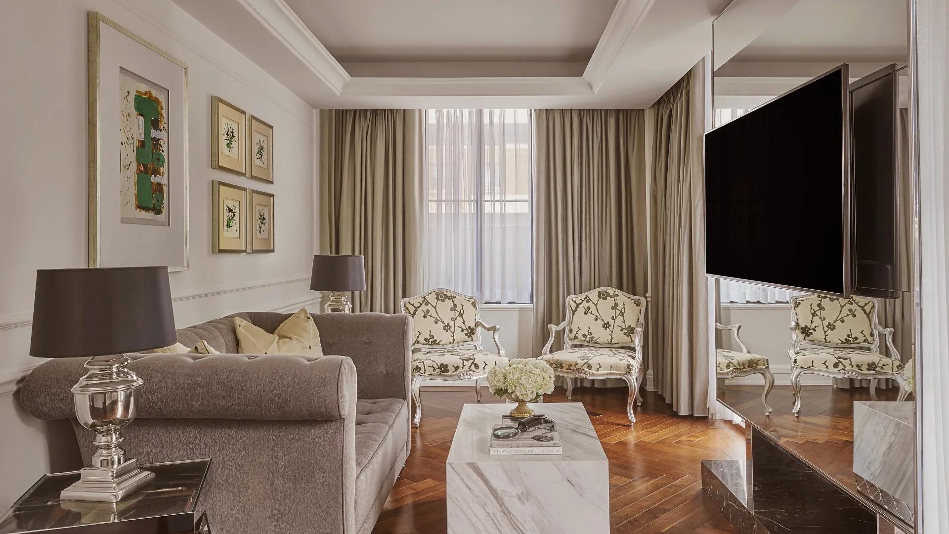 Spacious living room with a grey sofa, marble coffee table, two printed chairs, and a television hanging on a mirror wall.  