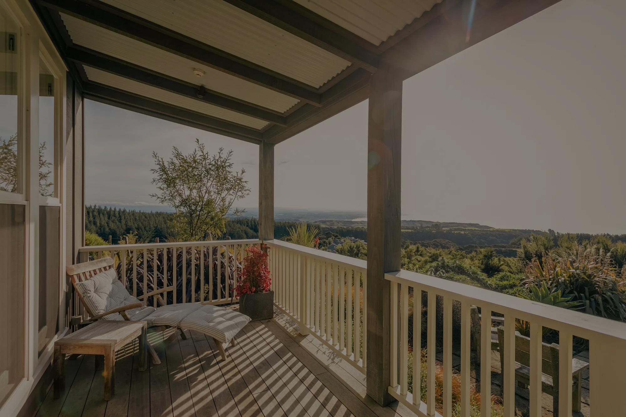 Covered balcony with a white railing and lounge chairs overlooking forested hills and ocean.