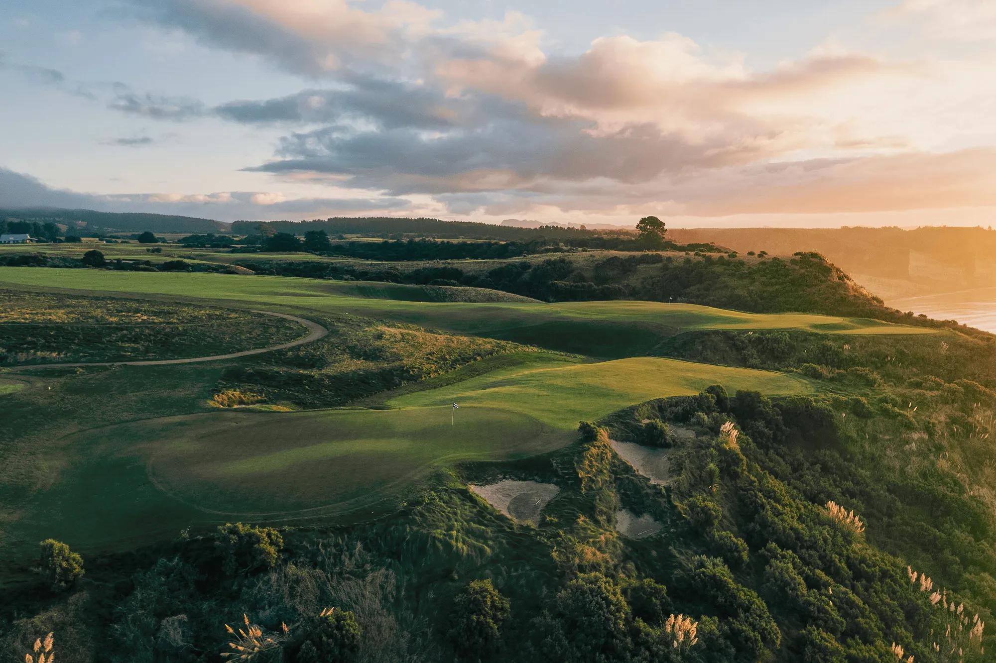 Aerial view of fariways perched along a cliffs edge, with pampas grasses and shrubs growing on the cliffside. 