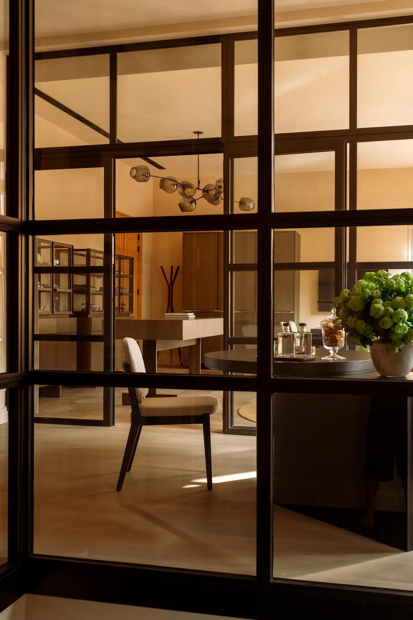 A view through iron crittal doors into a room with cabinets, desk and chair decorated with art, flowers and a chandelier