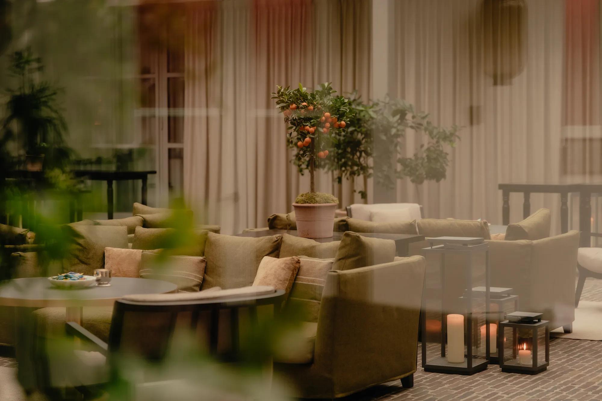 Dark green velevt sofas and coffee tables in a bright courtyard with brick flooring, deocrated with orange trees and large floor lanterns