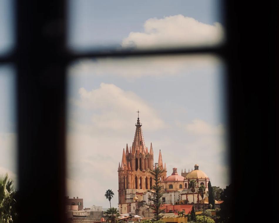 A window frame with wooden borders revealing a clear view of the Parroquia de San Miguel de Allende against the sky.