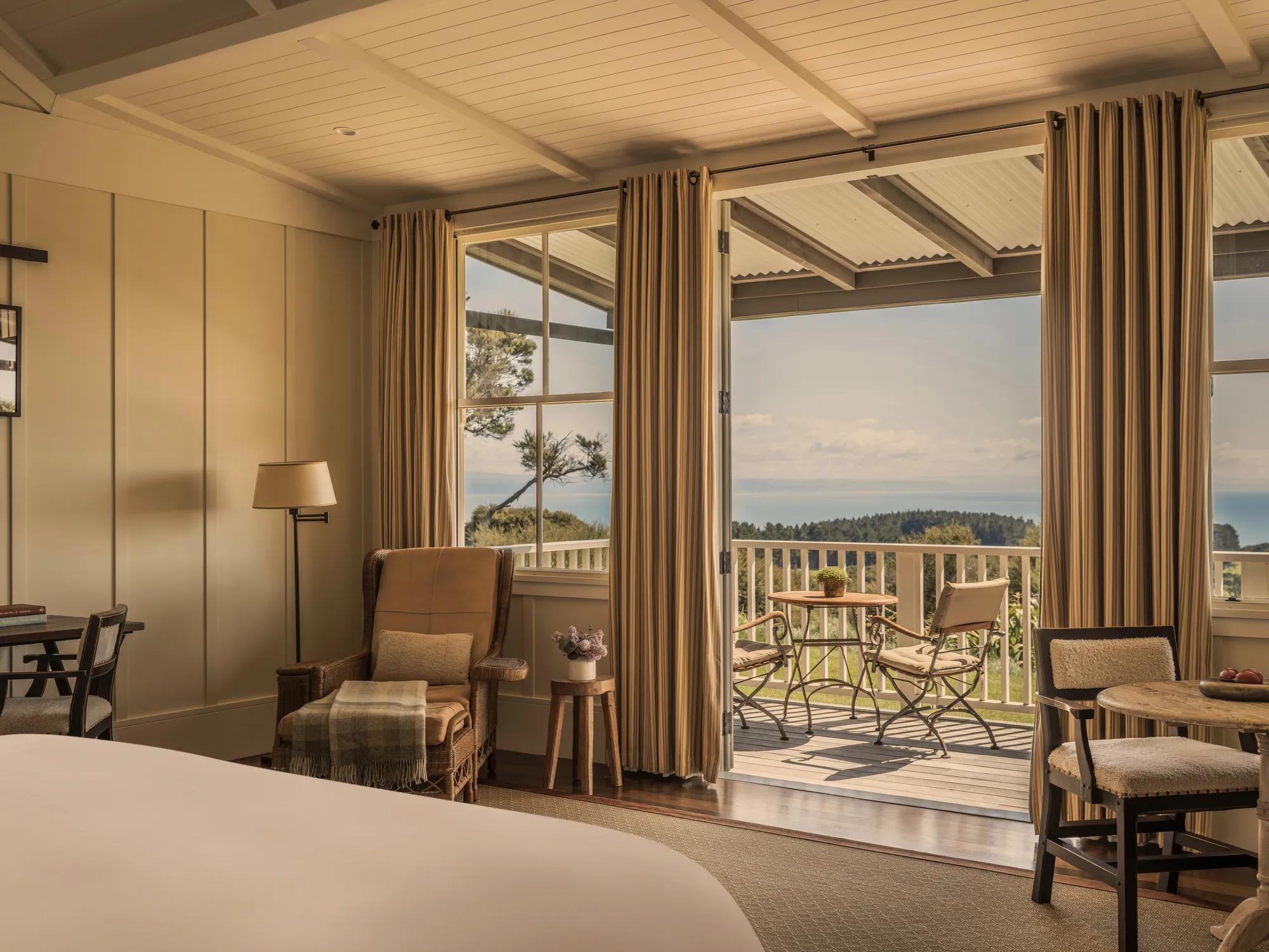 A hotel suite with neutral interiors, armchair, table with fresh fruit, desk, balcony and ocean views.