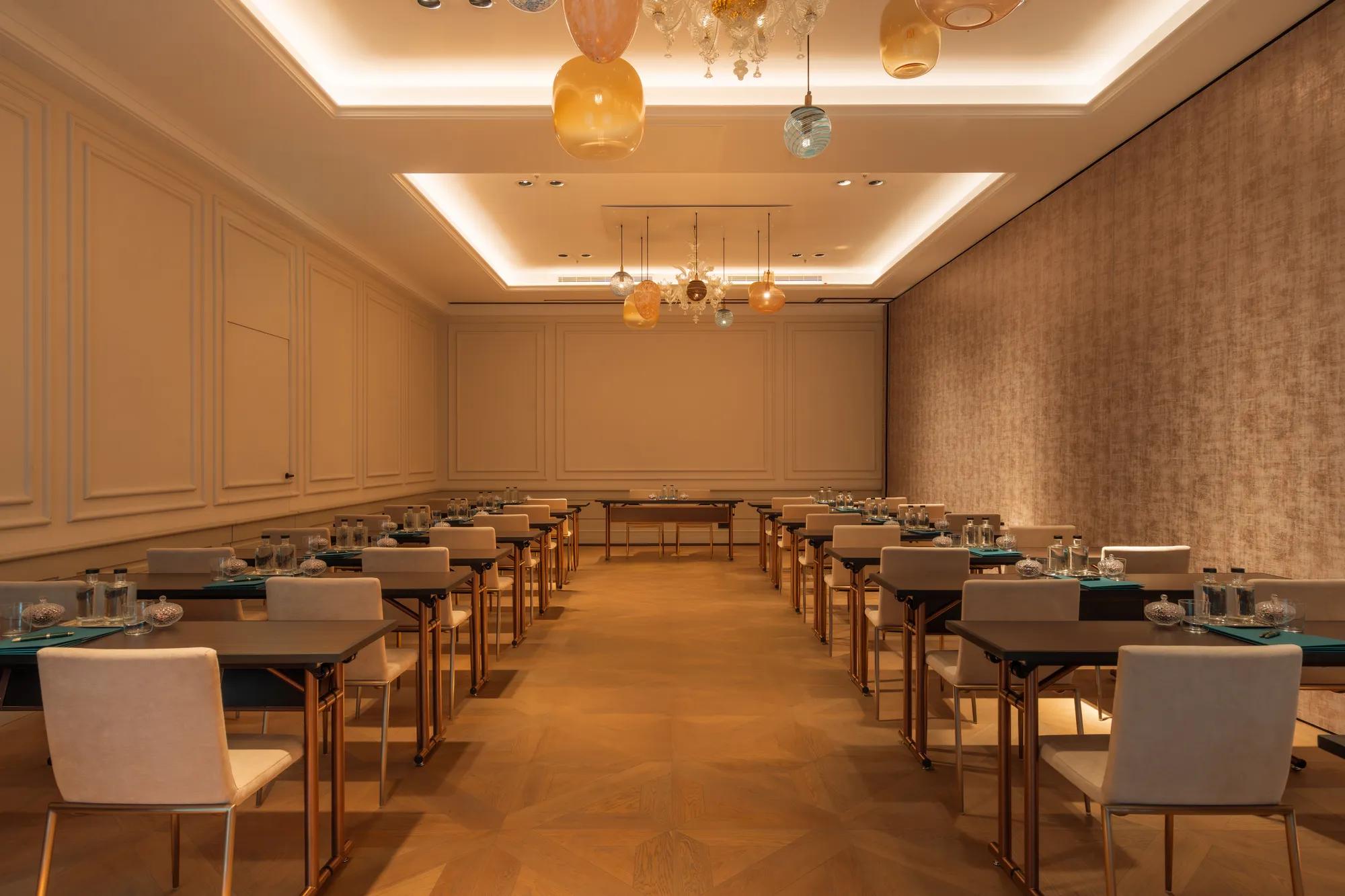A ballroom with chairs and tables set up in classroom style. The space has white wooden pannelling, wooden floors and glass chandeliers. 