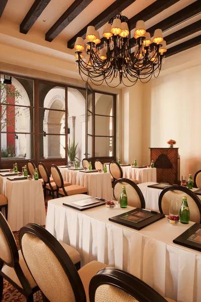 Meeting room with rectangular tables set for three, arranged in rows. Natural light enters from the side, with a chandelier overhead.