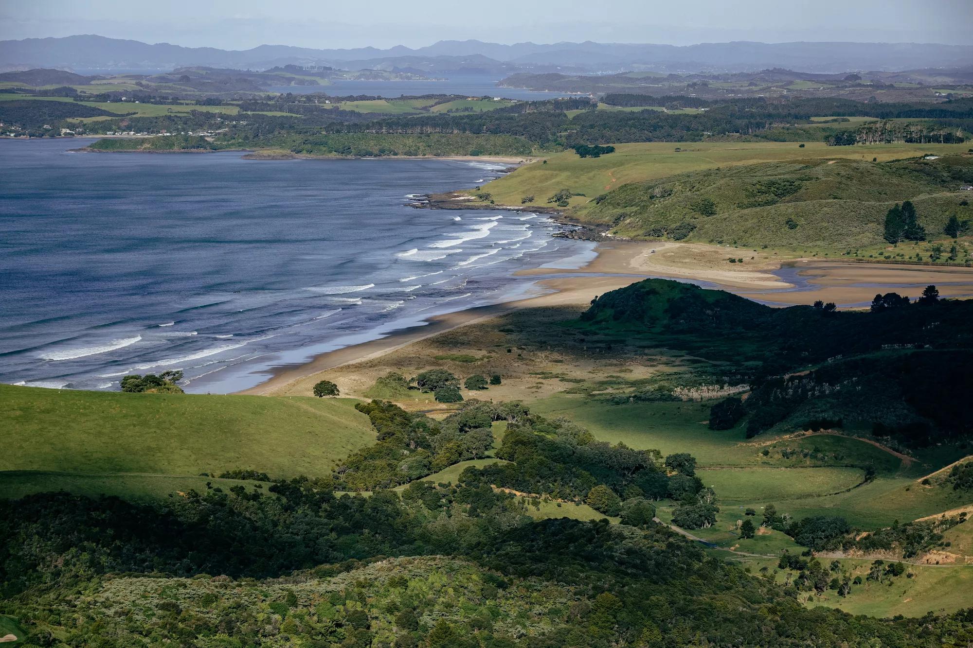 Aerial view of a beach with waves rolling in, the Bay of Islands are visible in the distance.