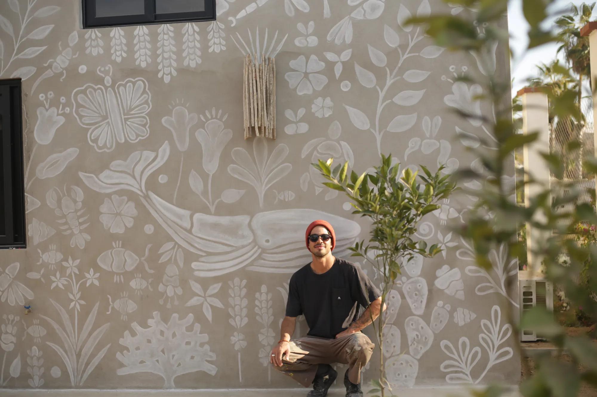 Rodrigo Azanza crouching beside a young plant in front of a hand-painted mural featuring a whale and botanical motifs, reflecting Baja-inspired art and nature.