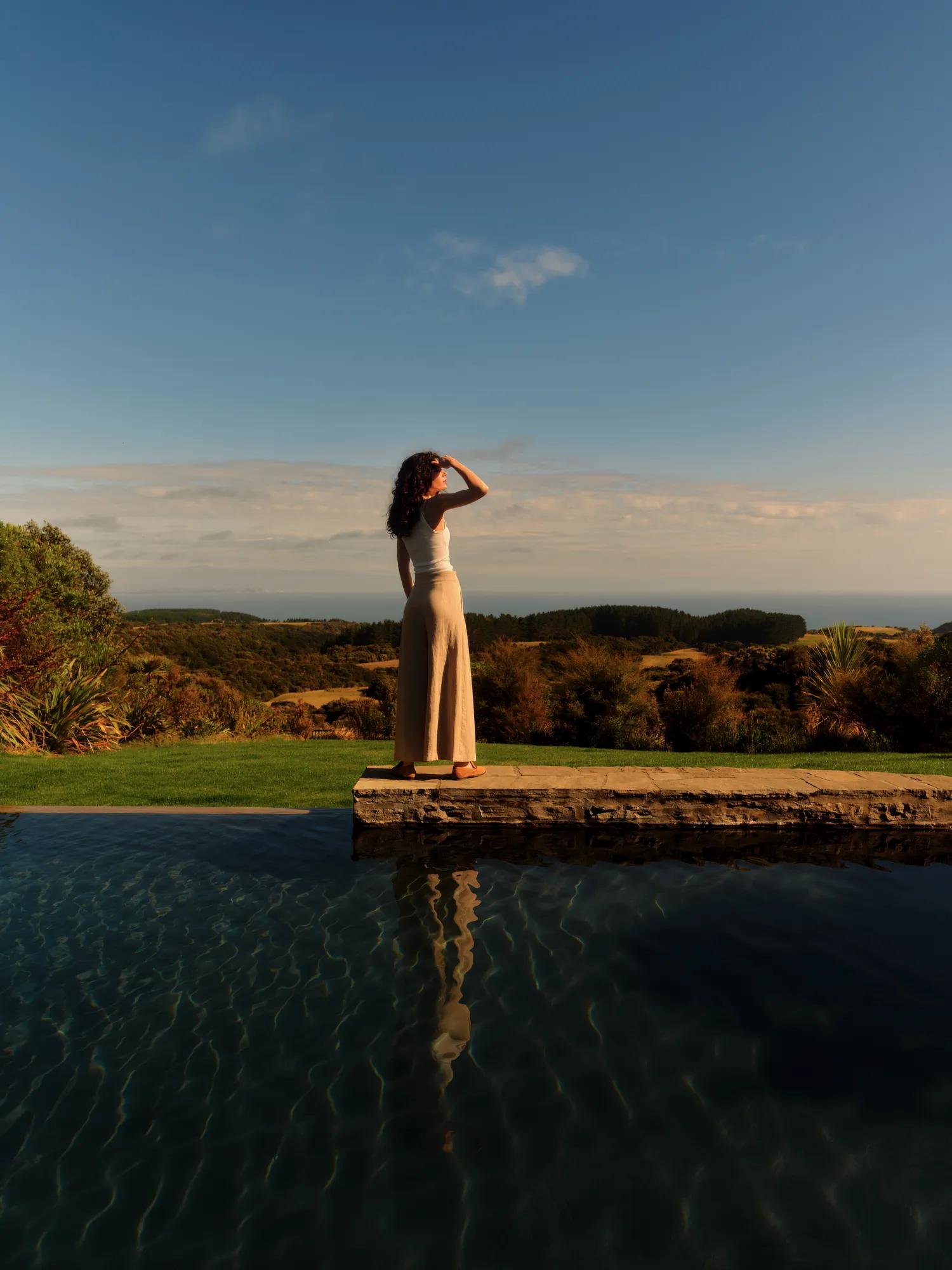 A woman standing beside an infinity pool overlooking the coastal landscape and lawn.