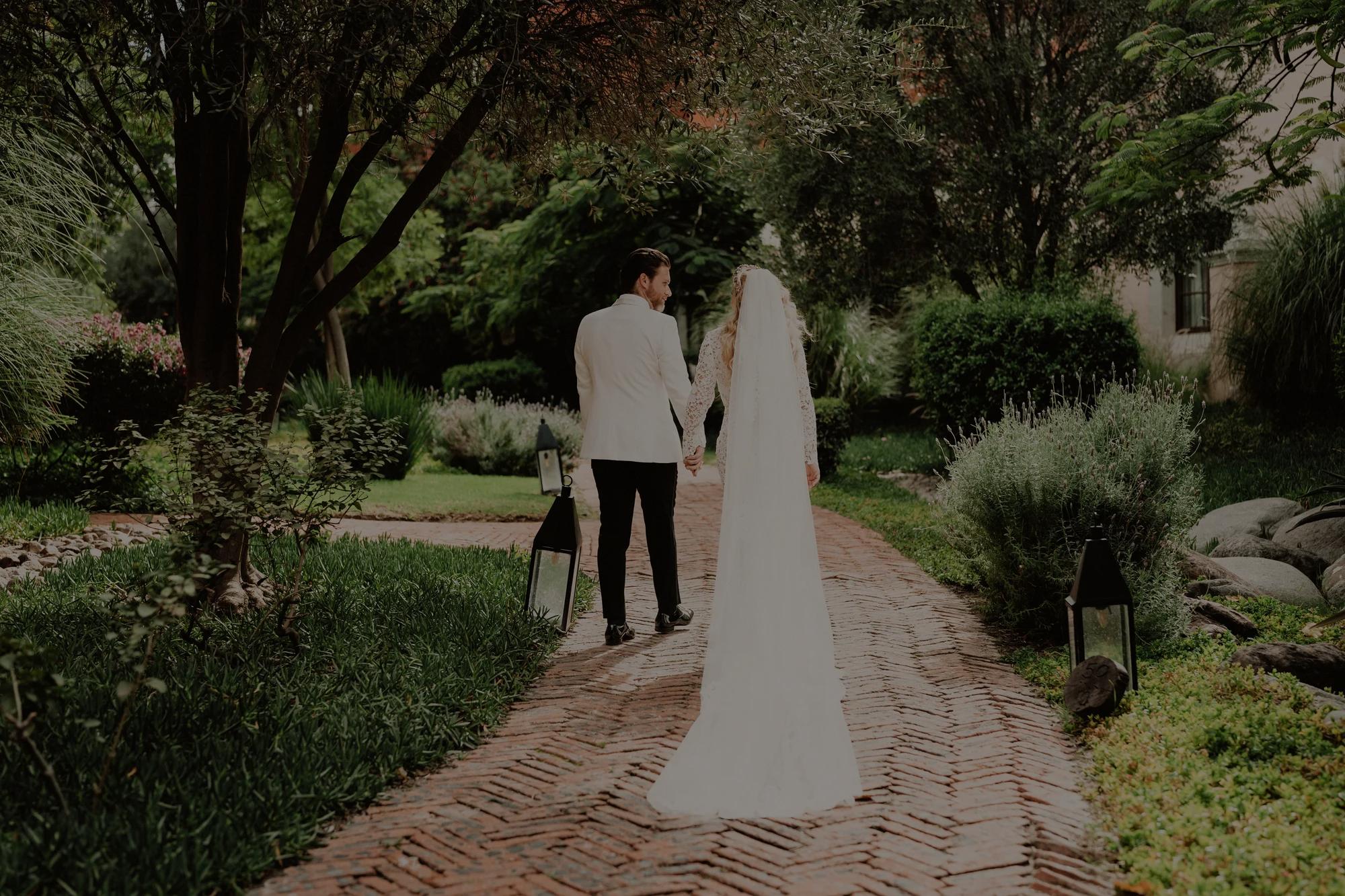 Bride and groom walking along a red brick path surrounded by lush greenery, sharing a quiet moment in a garden setting.