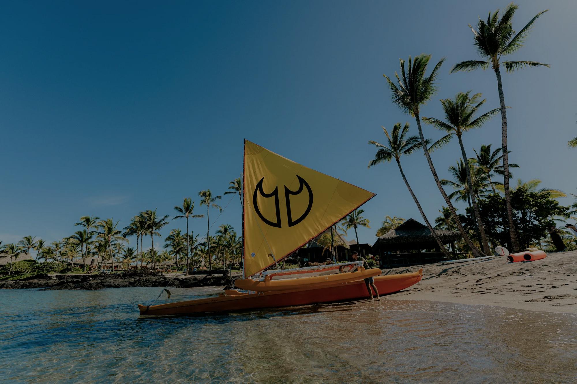 A sailing canoe with a bright yellow sail being pushed into the ocean off the shore of the beach with palm trees in the background.