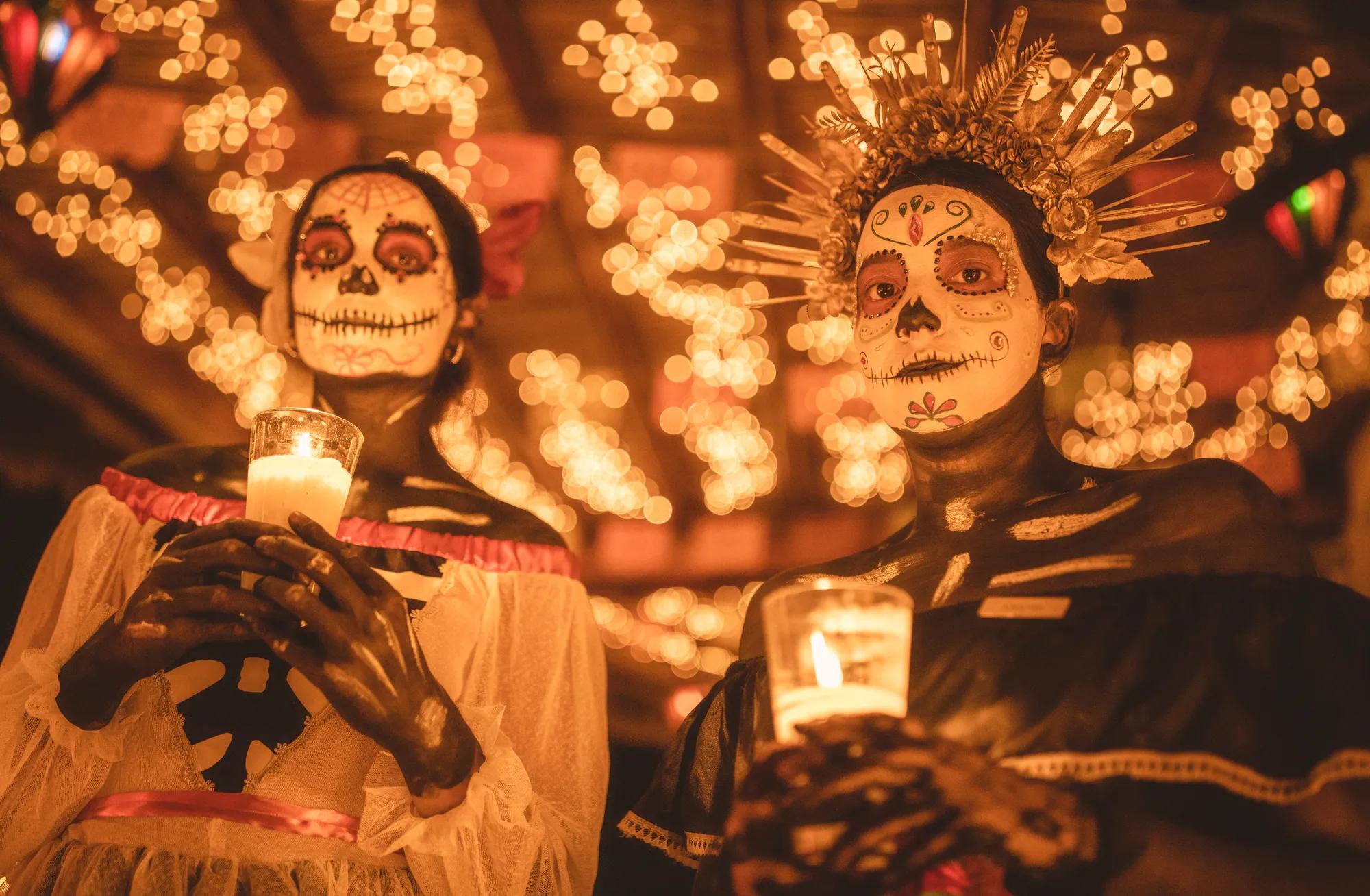 A woman in traditional folkloric dress dances gracefully, holding a decorative fan and lifting her lace skirt, under glowing golden lights. A man in a white outfit and hat stands smiling in the background, creating a festive Día de Muertos atmosphere.