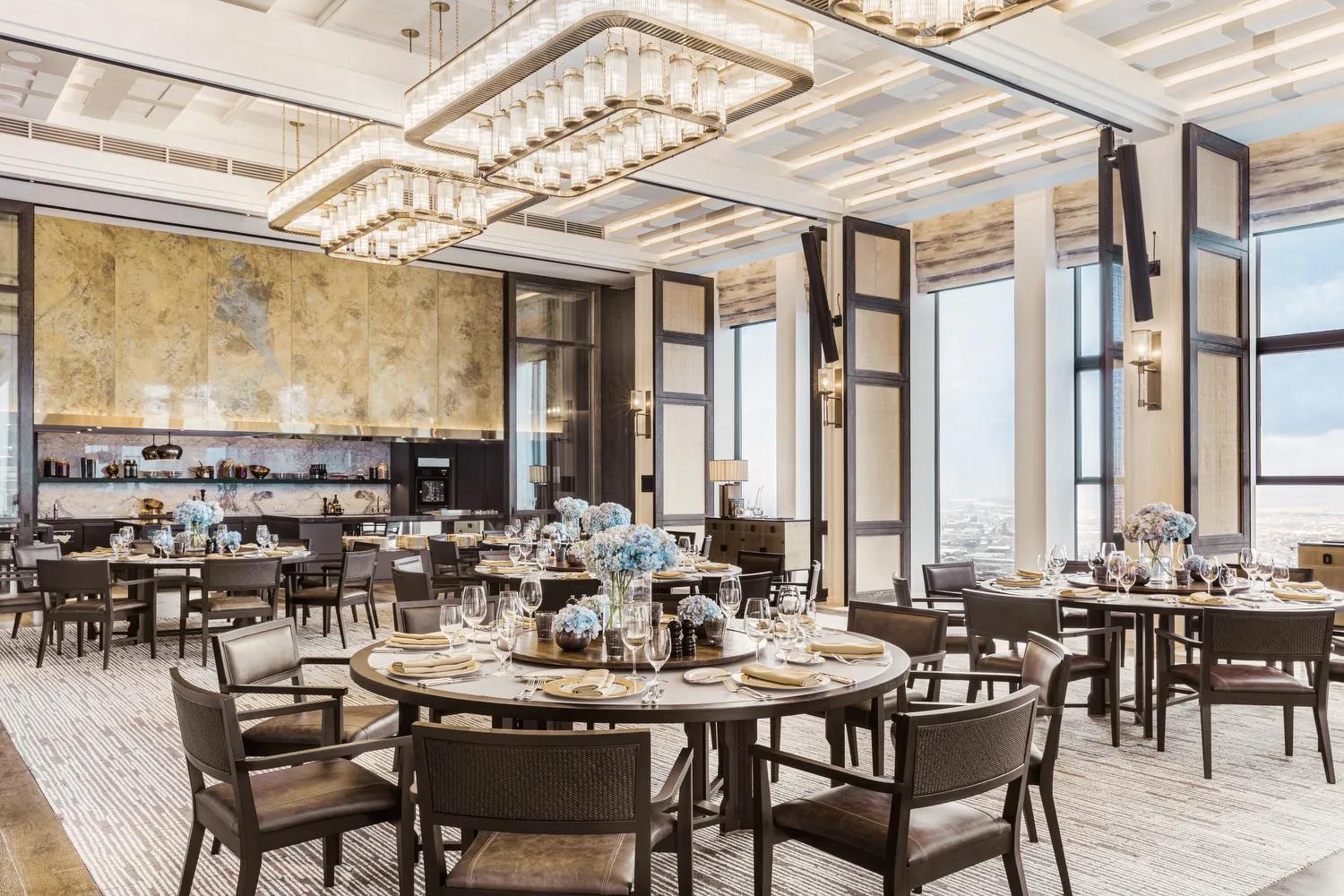 A modern dining area with round tables set for a meal, featuring blue floral centerpieces, elegant tableware, large windows, and contemporary chandeliers with a bar area in the background.