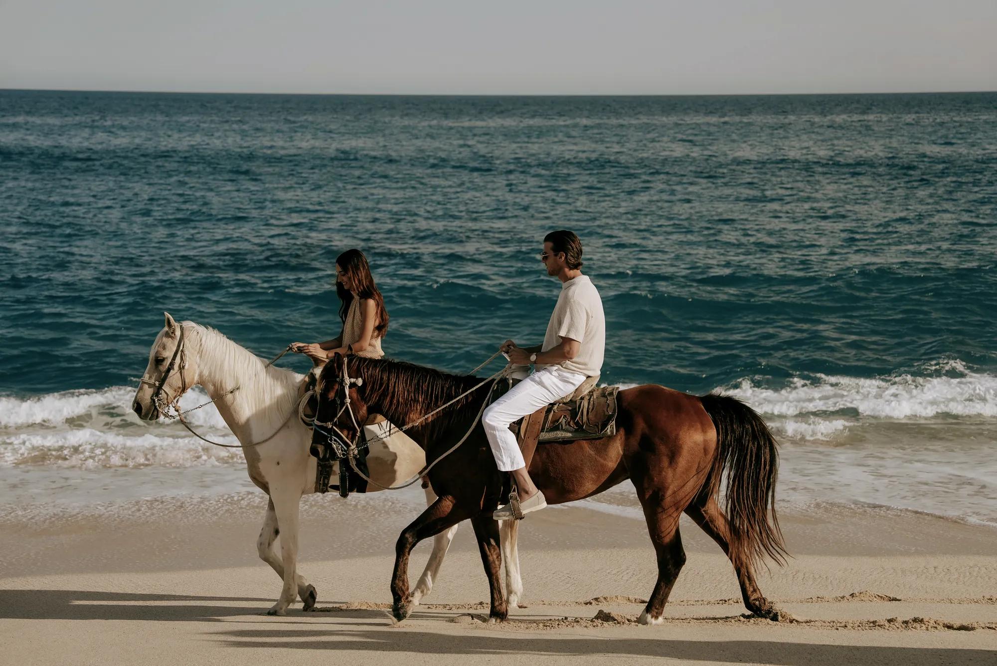 A couple riding horses along a sandy beach at sunset, waves gently rolling in beside them.