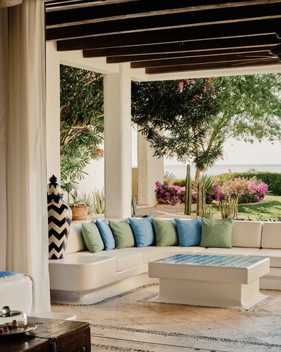 A bright suite with a bed and decorative headboard beside an open-air lounge area, featuring a built-in sofa with blue and green cushions, a tiled coffee table, and views of a garden with bougainvillea and cacti.