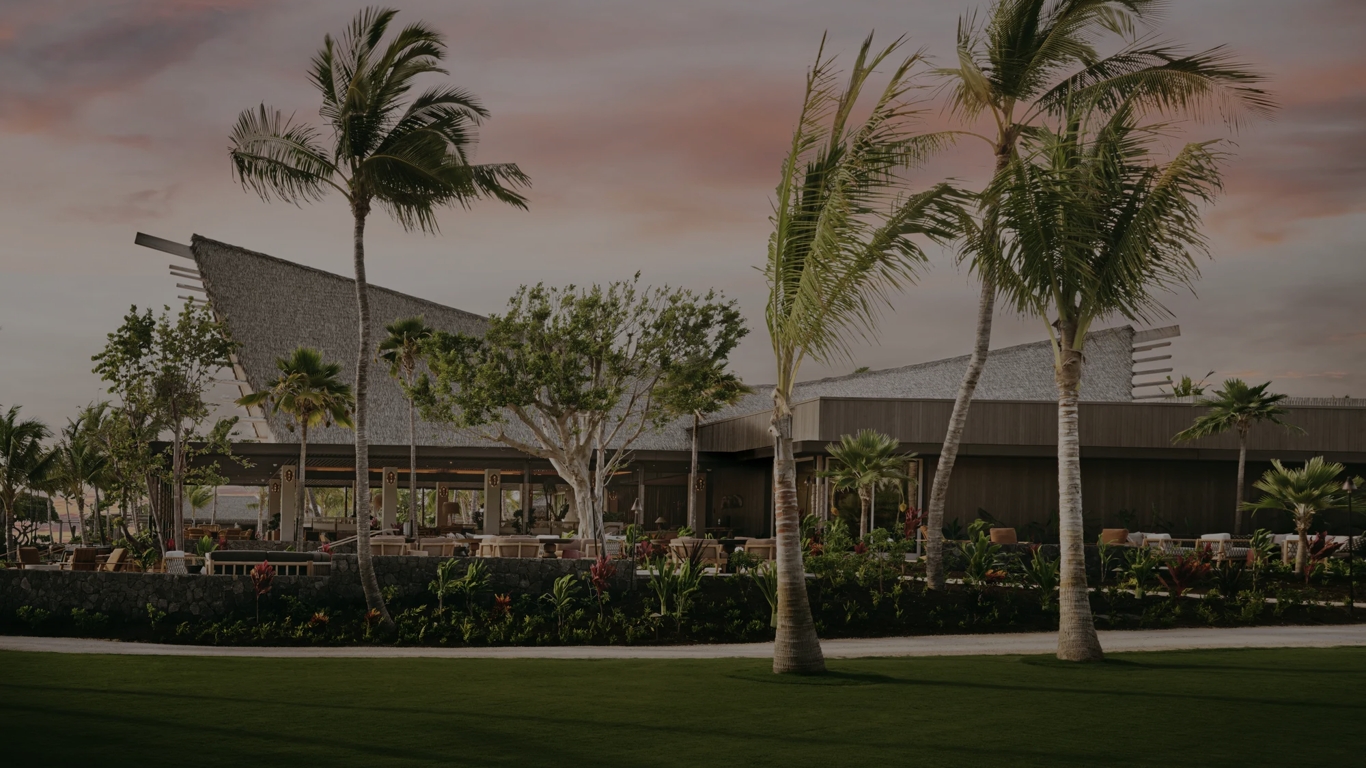 An A-frame restaurant exterior amongst palm trees at sunset.